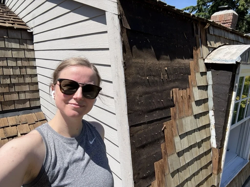 A woman wearing sunglasses and a sleeveless Nike athletic shirt taking a selfie outside on a sunny day. She is standing next to a house with white siding on one side and a partially repaired brown shingled roof on the other side.