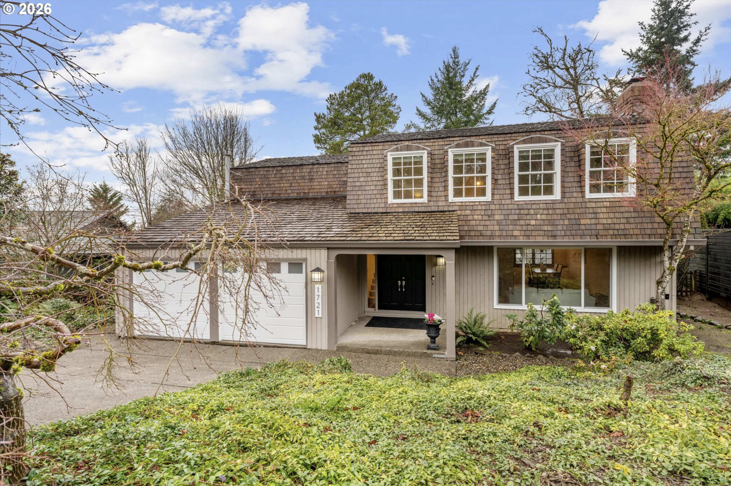 Front view of a two-story house with a shingled roof, white window frames, a garage door, and a front door. The house is surrounded by leafless trees and green bushes, with a driveway and a small garden area. The sky is blue with some clouds.