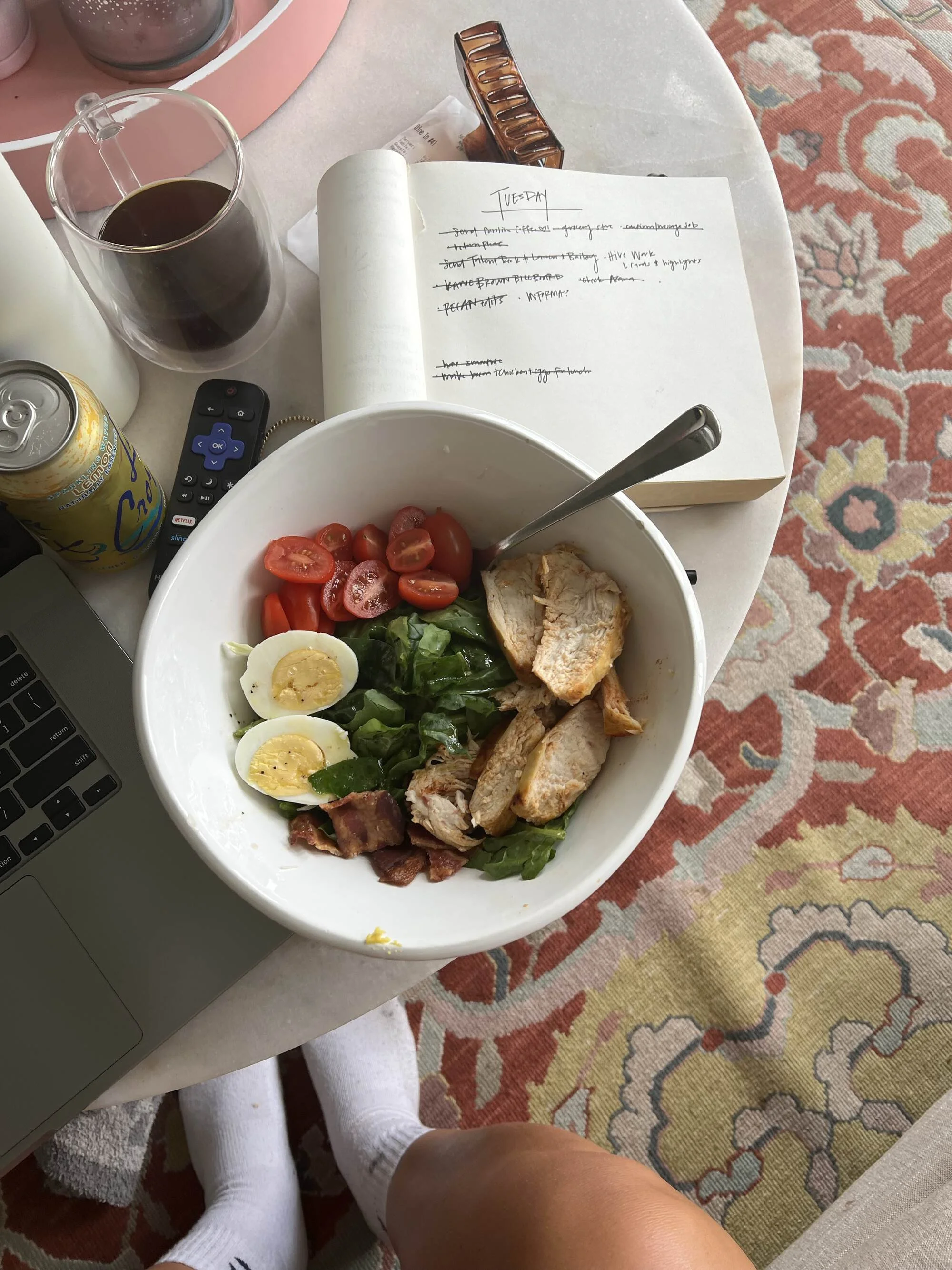 A table with a glass of dark soda, a can of sparkling water, a TV remote, a notebook with handwritten notes, a hair clip, a laptop, and a bowl of salad with cherry tomatoes, hard-boiled eggs, and grilled chicken, with a person sitting nearby wearing white socks and a blanket on their lap, all on a patterned rug.