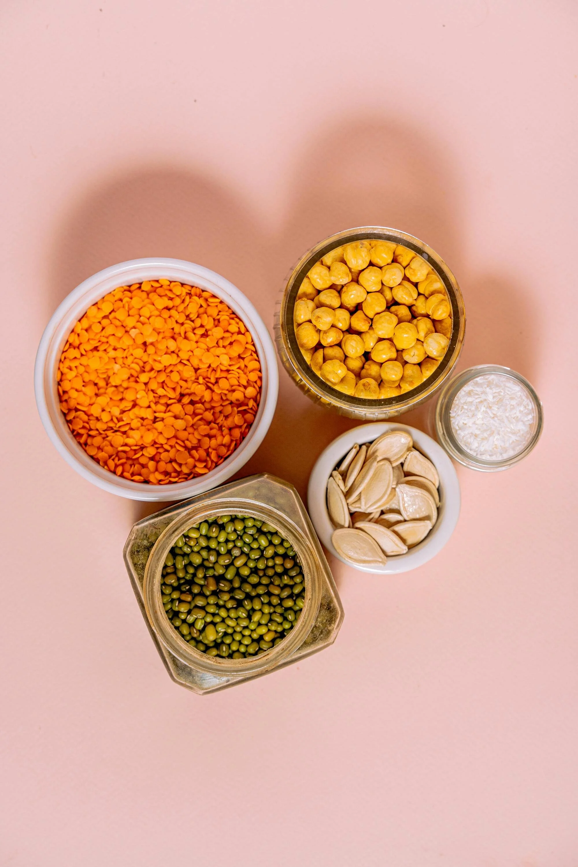 Top view of various containers with different types of legumes and seeds on a pink surface.
