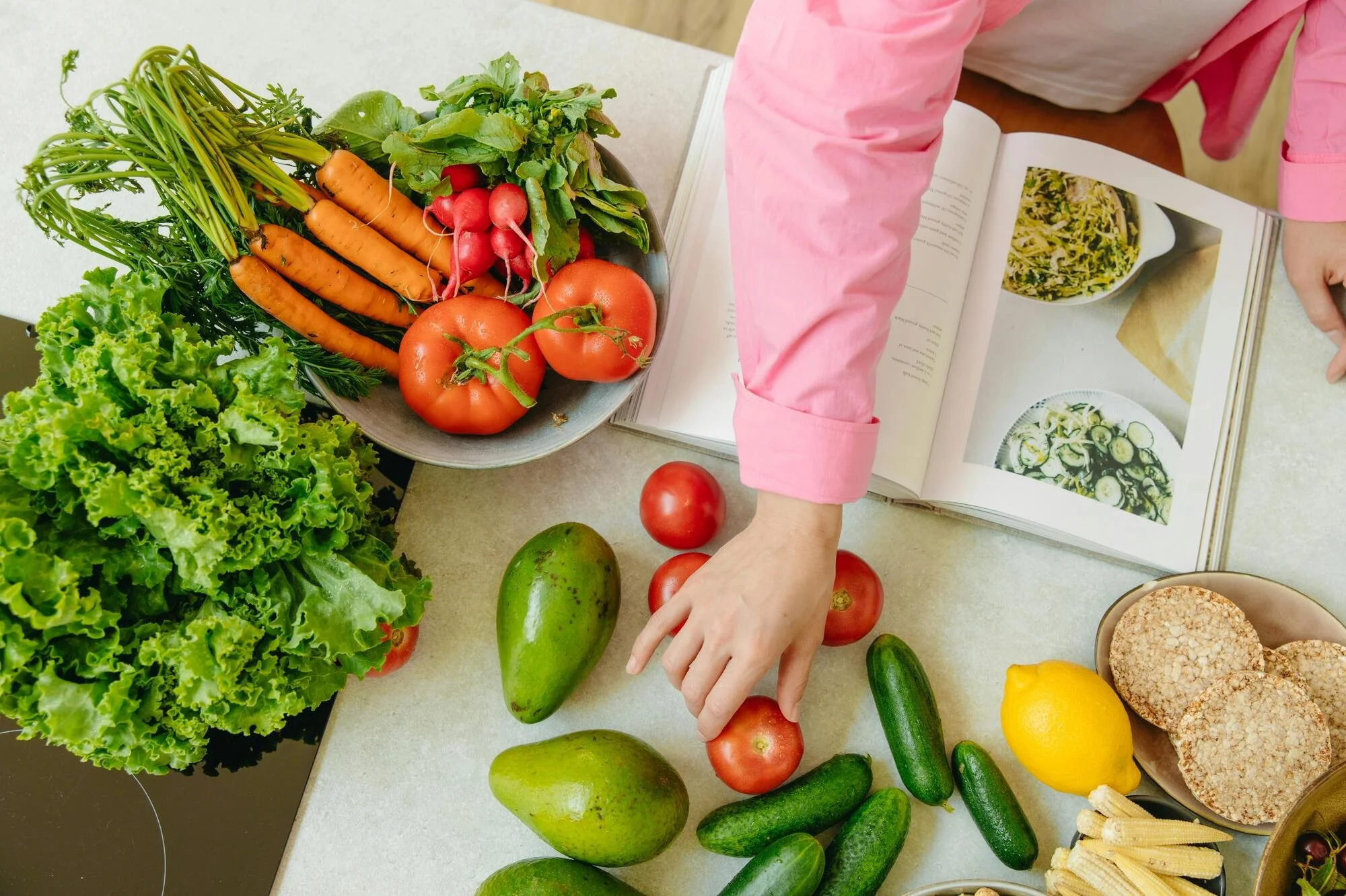 A person wearing a pink shirt is holding a tomato and placing it on a table with various vegetables, including carrots, radishes, cucumbers, two tomatoes, an avocado, a yellow bell pepper, and green leafy lettuce and herbs, with a cookbook open nearby.