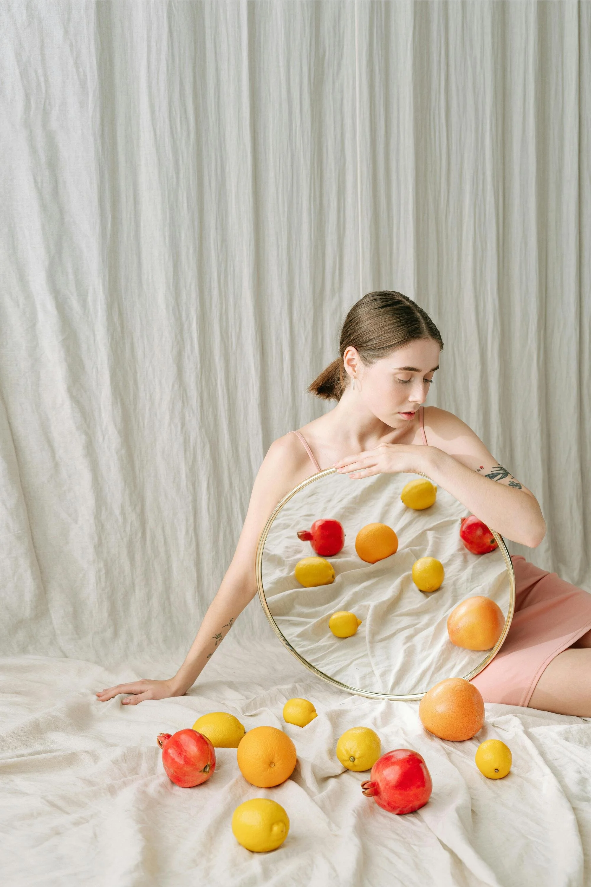 A young woman with brown hair wearing a pink dress sitting on the floor, holding a large circular mirror that reflects a variety of bright fruits, including pomegranates, yellow lemons, and oranges, with a cream-colored curtain in the background.