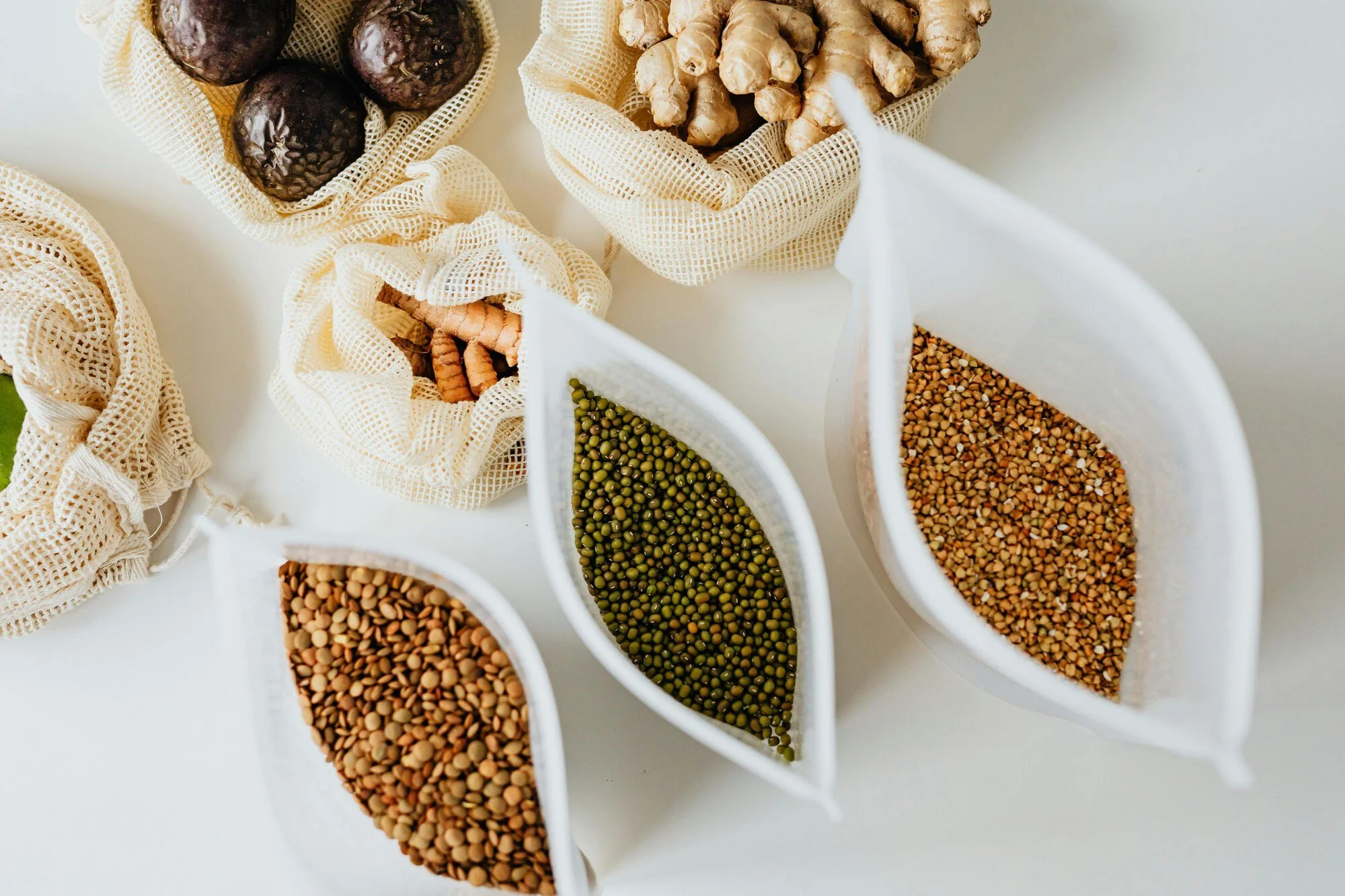Various spices stored in white mesh pouches and containers on a white surface, including dried ginger roots, black garlic, green lentils, buckwheat, and coriander seeds.