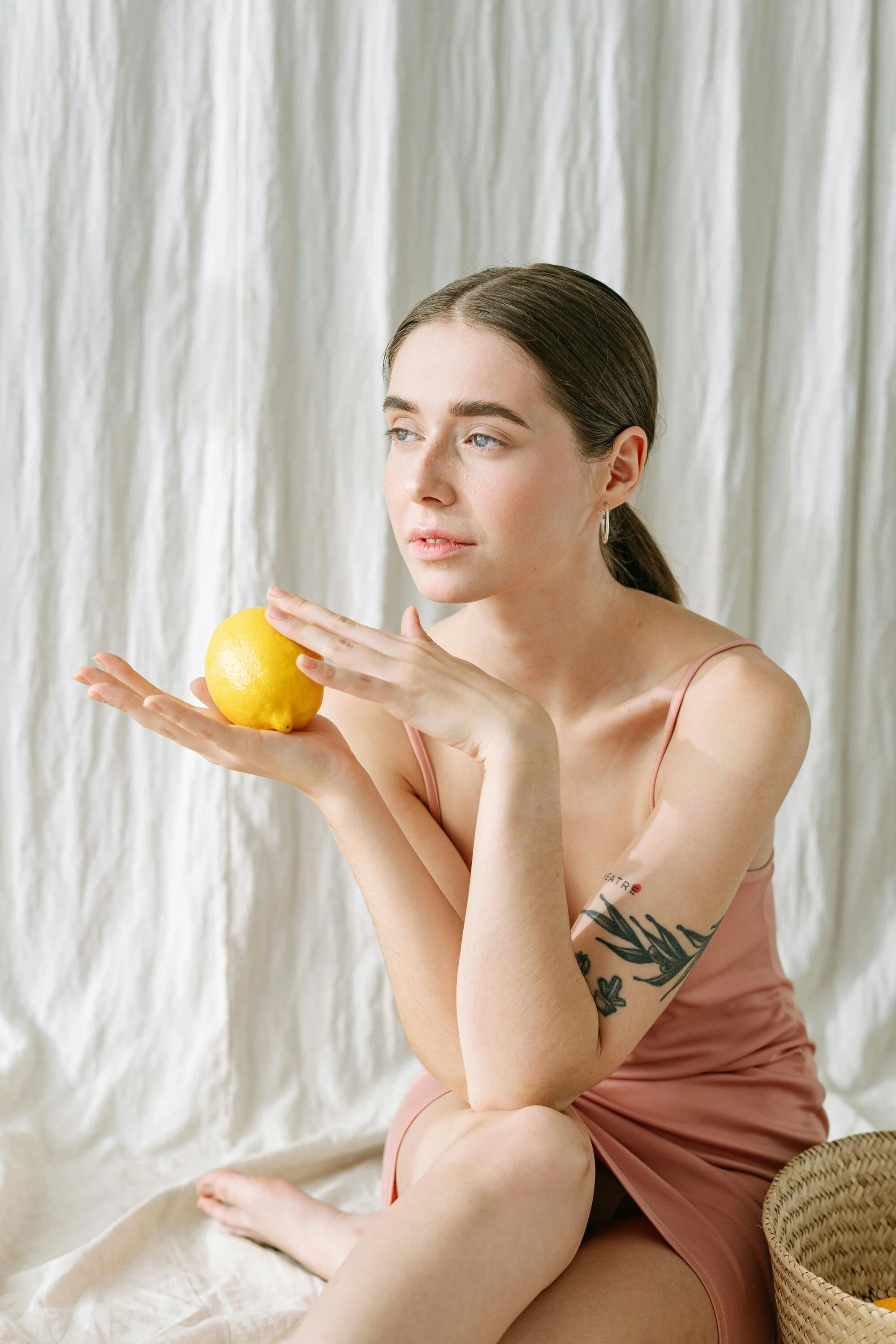 A young woman with braided brown hair and tattoos on her left arm sits on a beige fabric with a yellow lemon in her left hand.