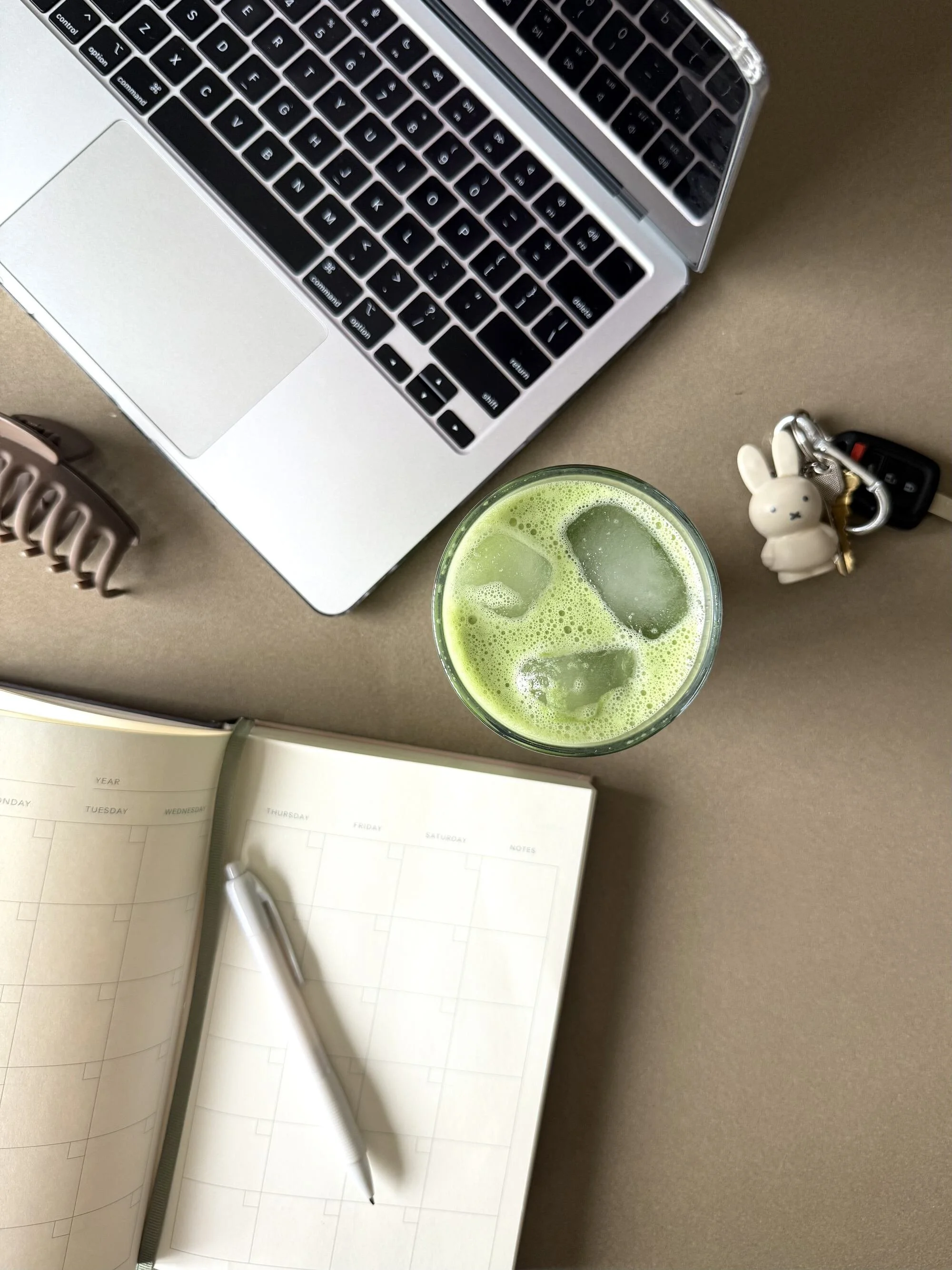 A workspace with a laptop, a glass of green drink with ice, a small bunny keychain, a set of keys, a planner, and a white pen.
