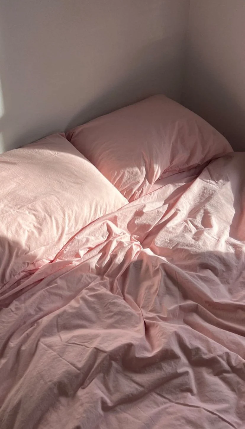 A bed with light pink sheets and pillows, slightly rumpled and unmade, in a corner of a room with a plain wall.