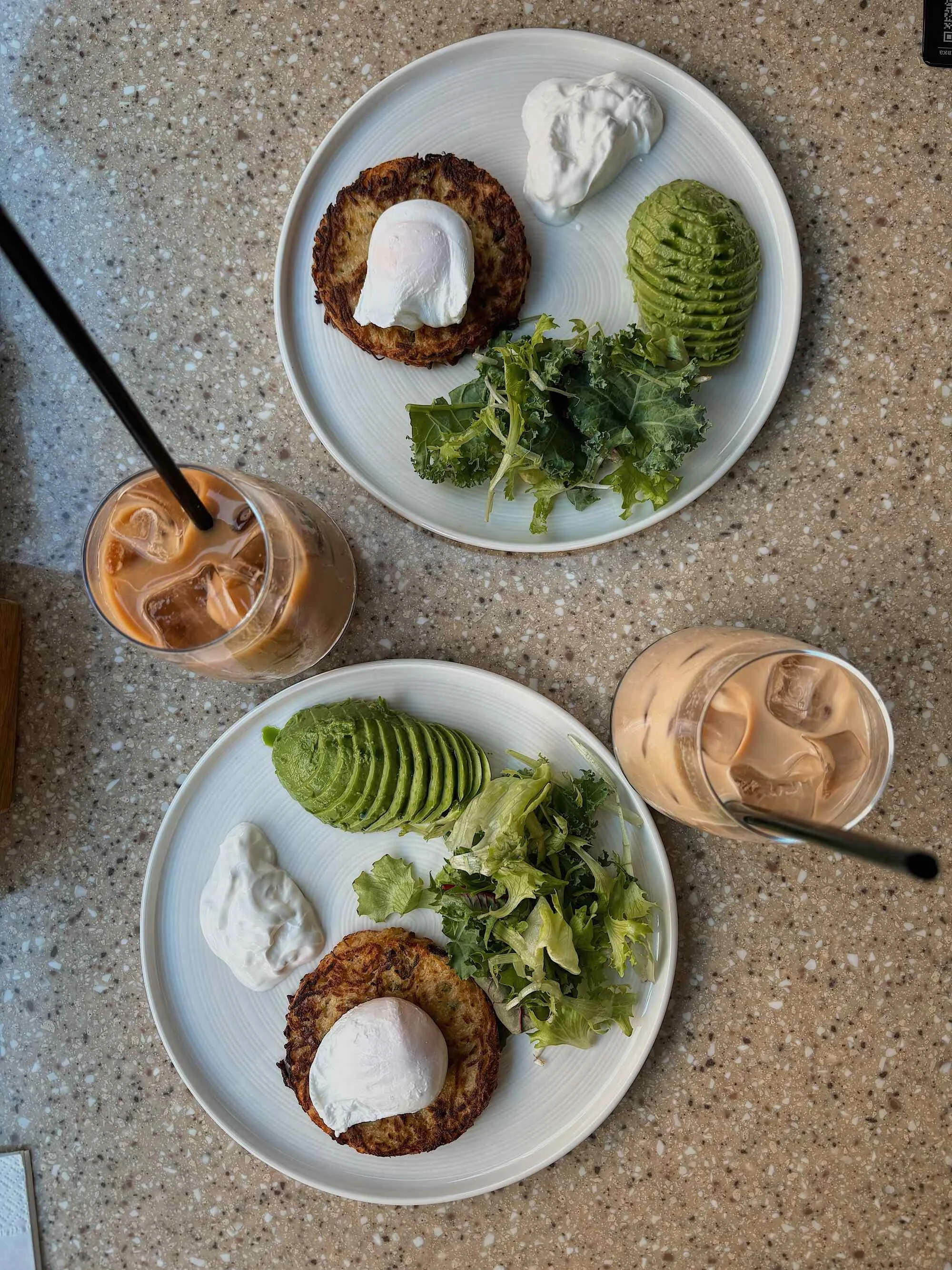 Two plates with hash browns topped with sour cream and a poached egg, sliced avocado, mixed greens, and dollops of sour cream, with iced coffee drinks nearby on a speckled countertop.