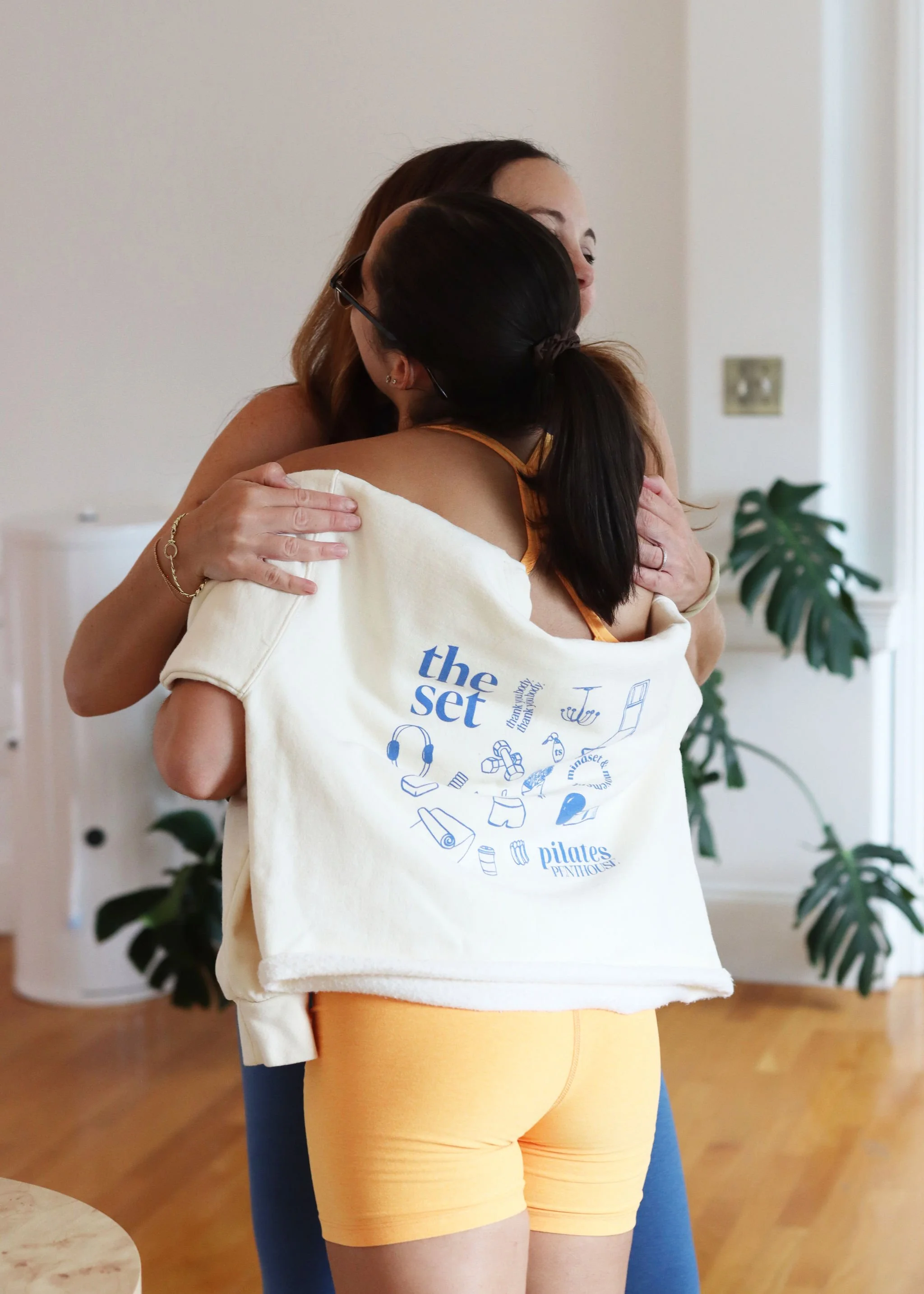 Two women hugging each other indoors, with one wearing a yellow top and yellow shorts, and the other wearing a white sweatshirt with blue graphics, in a room with wooden floors and a potted plant.