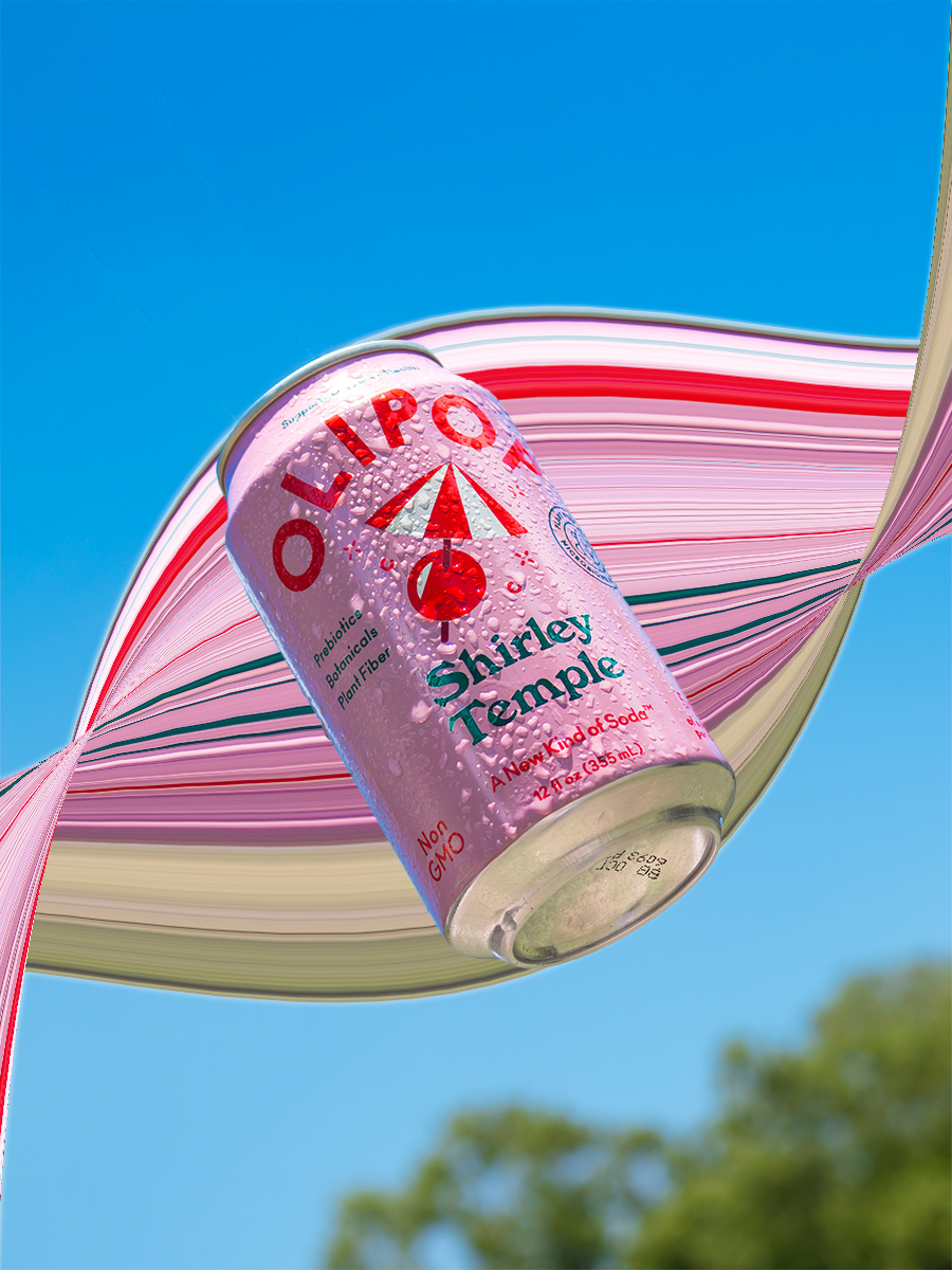 A pink can of Shirley Temple soda tilted against a pink, white, and green-striped parasol, with a bright blue sky and some trees in the background.
