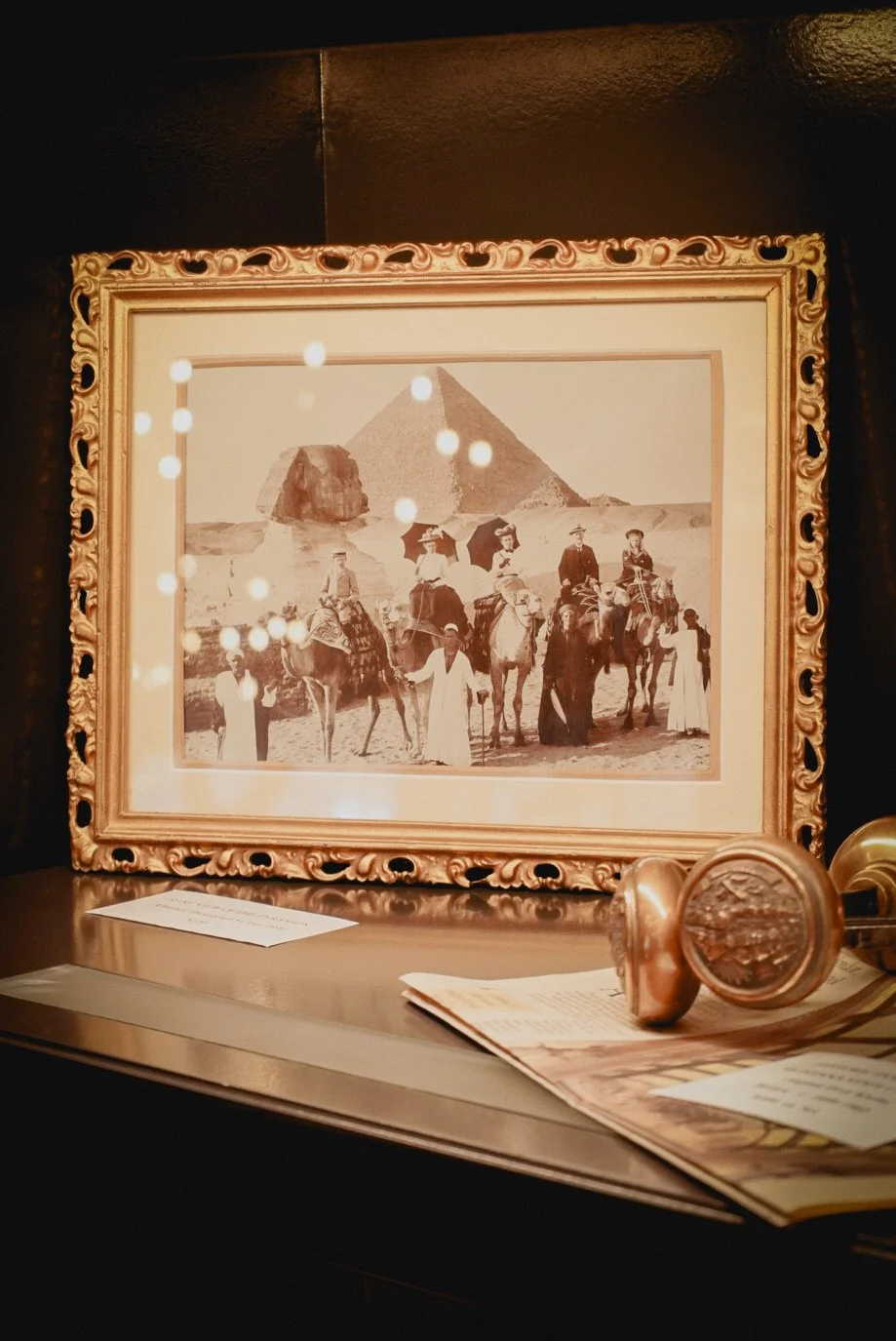 A framed black-and-white photograph of a group of people with camels in front of the Pyramids of Giza, displayed inside a glass case with small spotlights overhead. There are some documents and a wax seal on the surface below the picture.