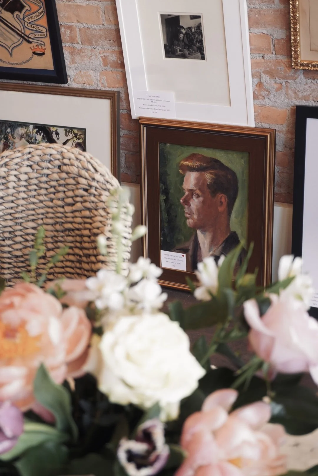 Framed portrait of a man with styled hair, set against a dark background, displayed on a brick wall among other framed artworks. Flowers and a woven chair in the foreground.