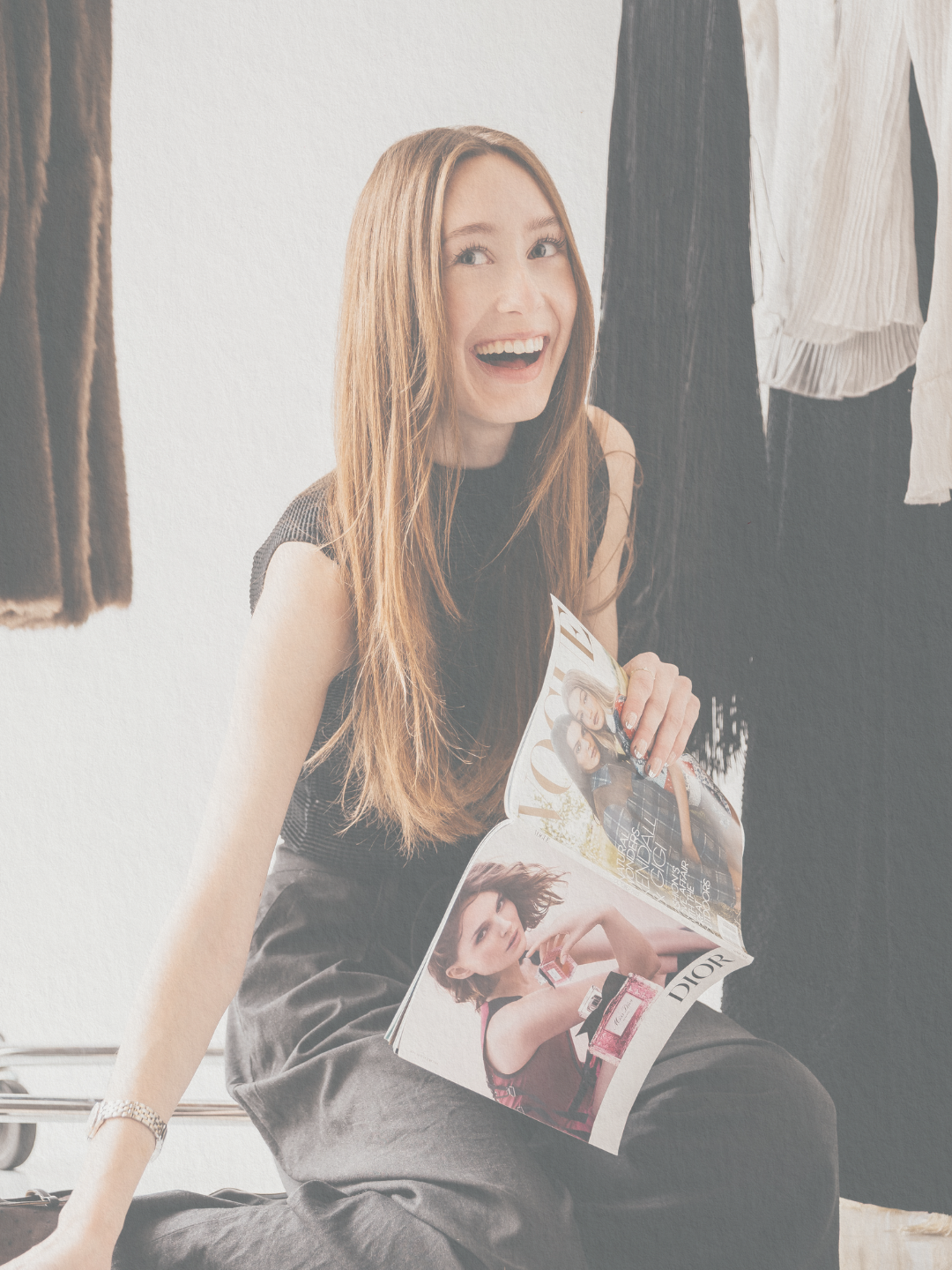 A woman with long red hair sitting on a bench surrounded by hanging clothes, holding a fashion magazine, and smiling.