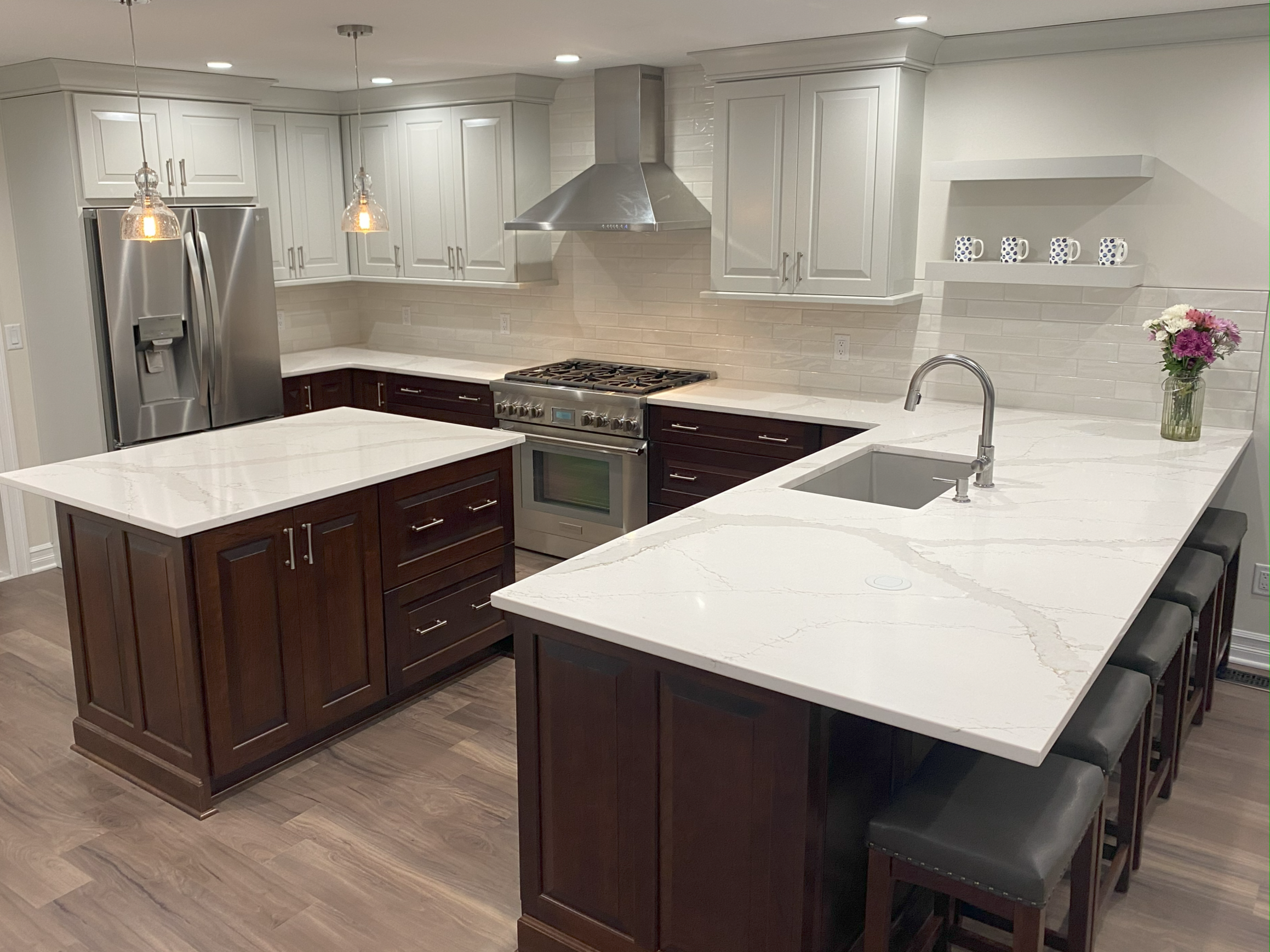 Modern kitchen with white and dark wood cabinets, white marble countertops, stainless steel appliances including a refrigerator and oven, and a kitchen island with bar stools. There are pendant lights above the island, and a vase with flowers on the counter.