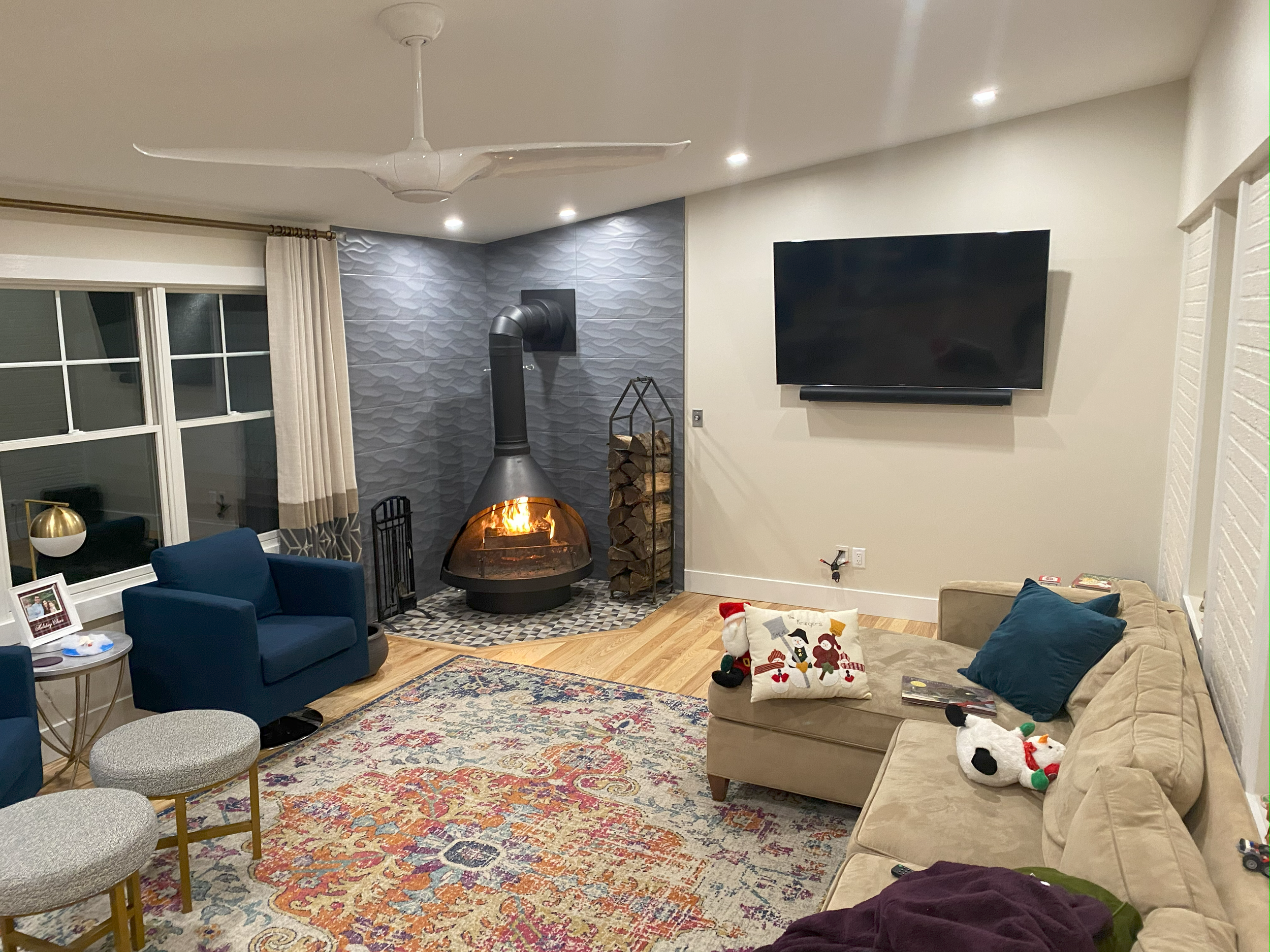 Living room with a wood-burning fireplace on a textured gray wall, a mounted flat-screen TV, a beige sofa with holiday-themed pillows, a blue armchair, small round stools, a colorful area rug, and Christmas plush toys on the sofa. A ceiling fan and window with curtains are also visible.