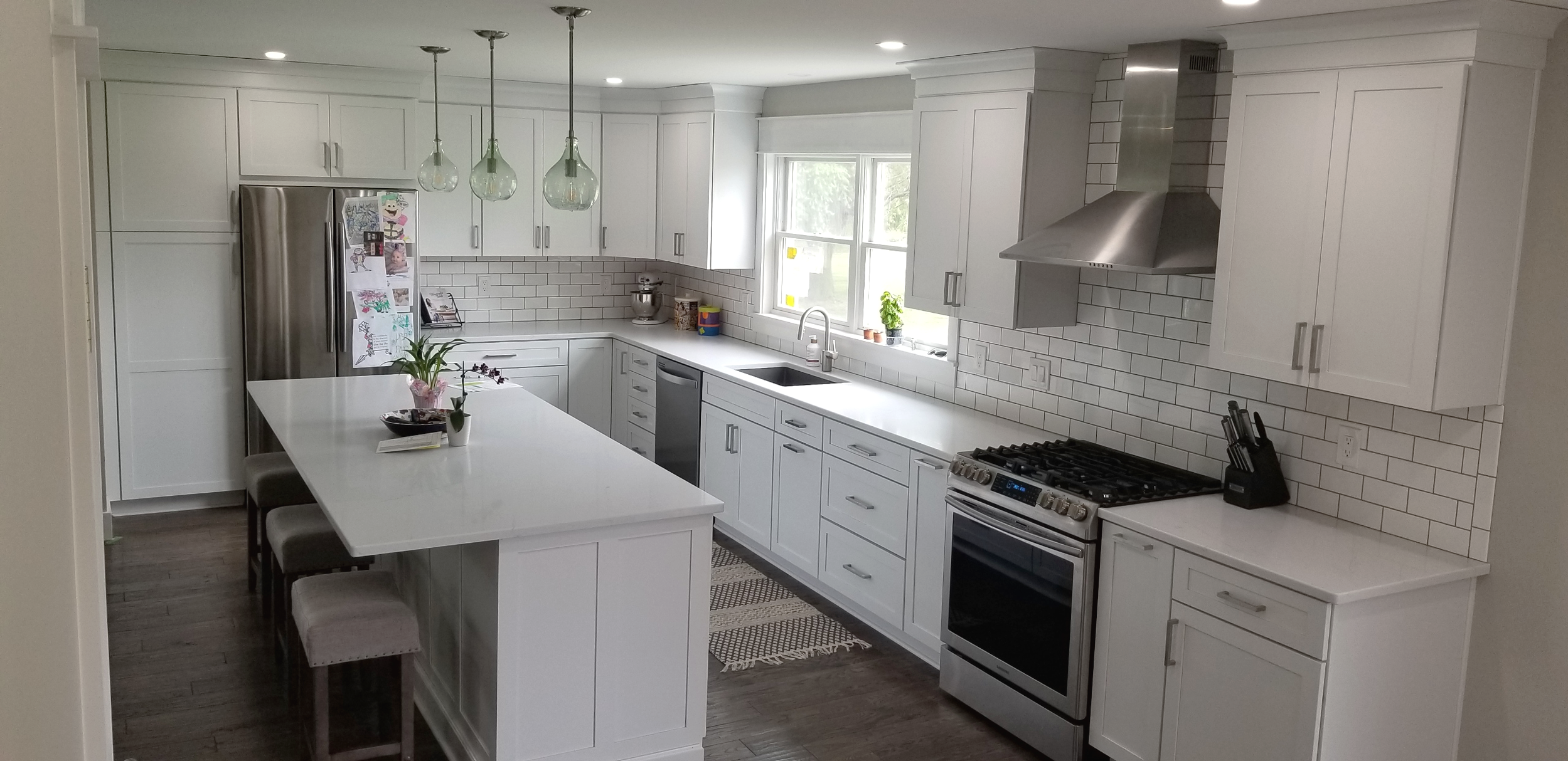 Modern kitchen with white cabinets, stainless steel appliances, subway tile backsplash, and a large island with bar stools, illuminated by three pendant lights.