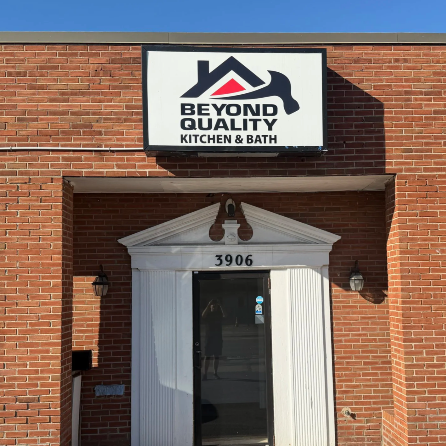 Brick building with a sign reading 'Beyond Quality Kitchen & Bath' featuring a logo of a house with a hammer. The building has the number 3906 above the entrance, which is a glass door surrounded by white decorative trim.