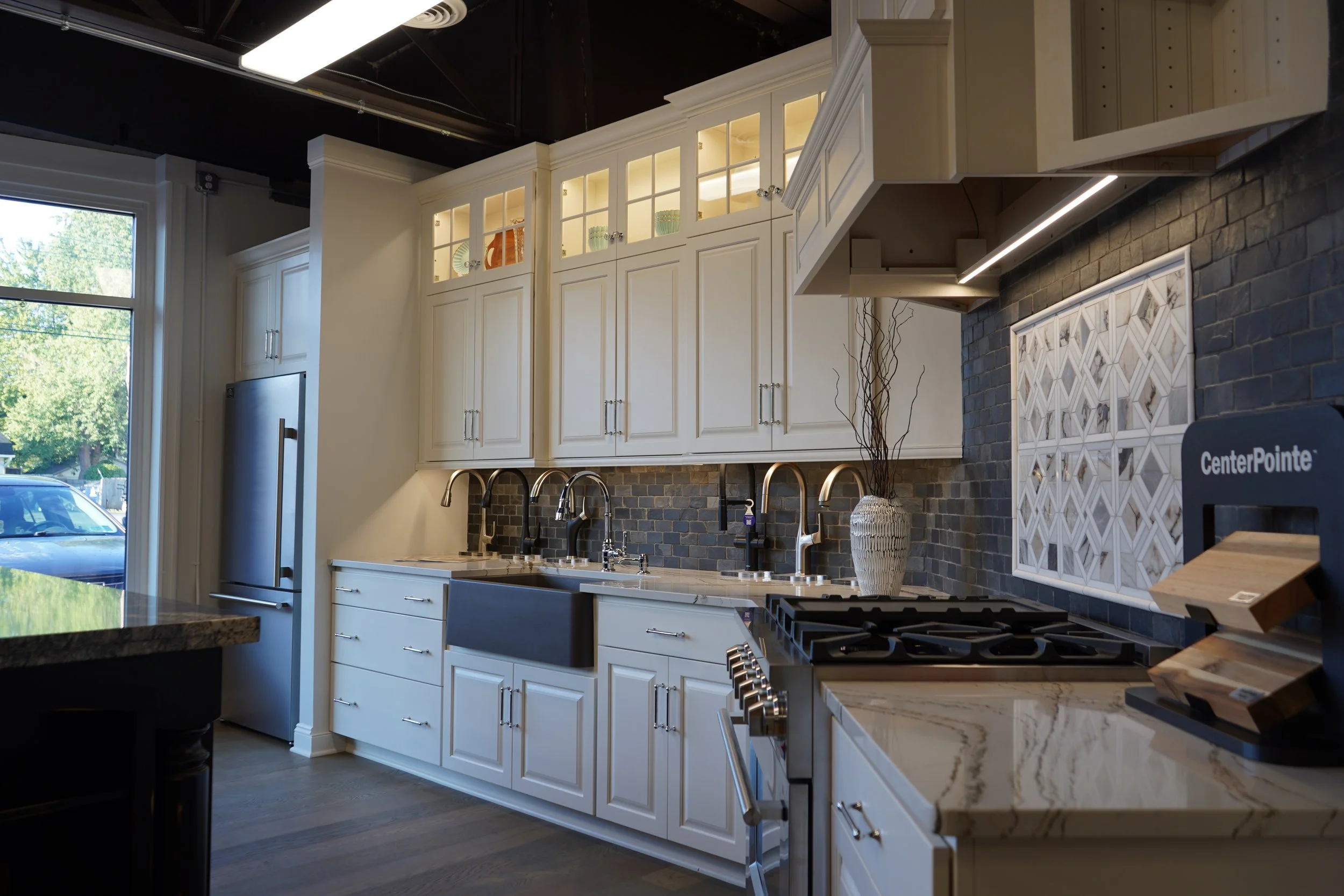 Modern kitchen with white cabinets, a black brick backsplash, a marble countertop, and a stainless steel refrigerator, with a window showing outdoor greenery.
