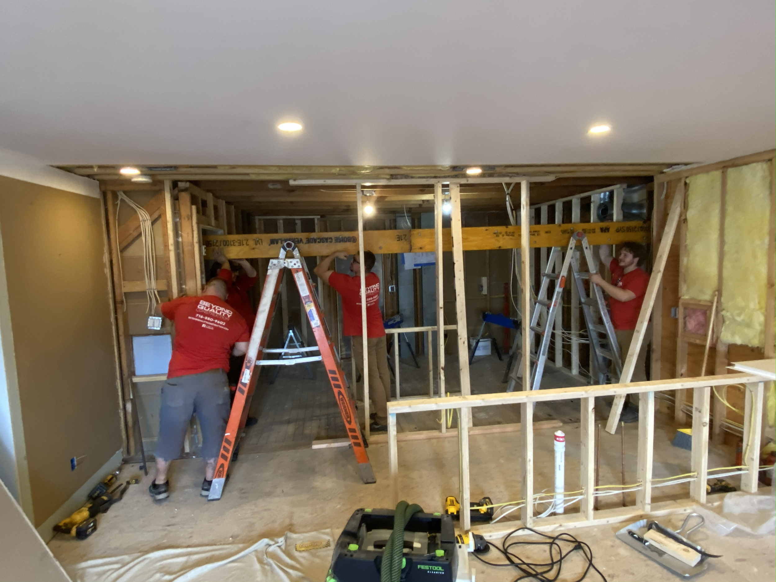 Three workers in red shirts installing a wall frame in a room under renovation. They are using ladders and tools, with exposed wooden studs and electrical wiring.