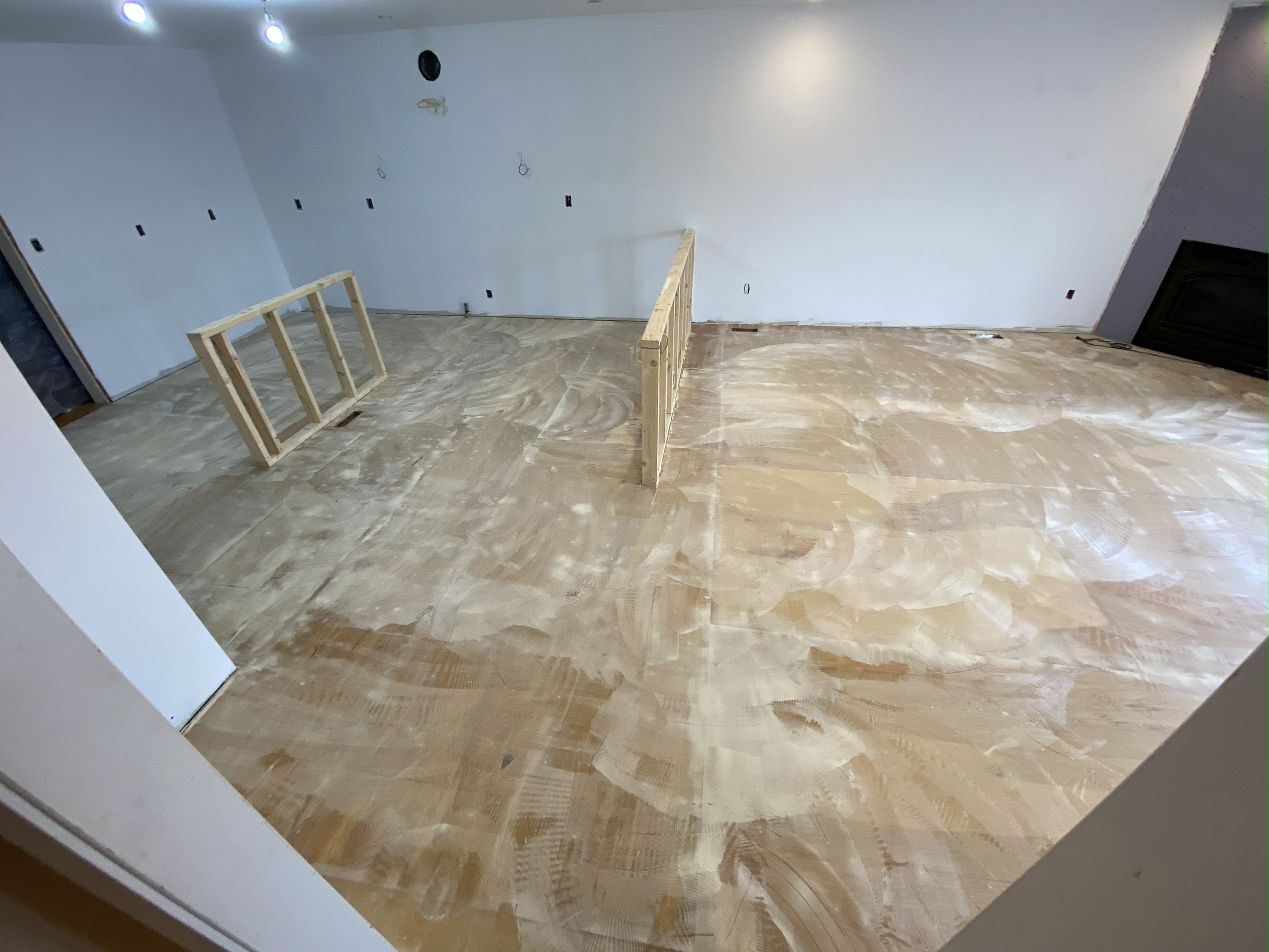 An unfinished room with a partly constructed wooden railing, drywall with electrical outlets on the walls, and a floor covered in visible joint compound or sanding residue.