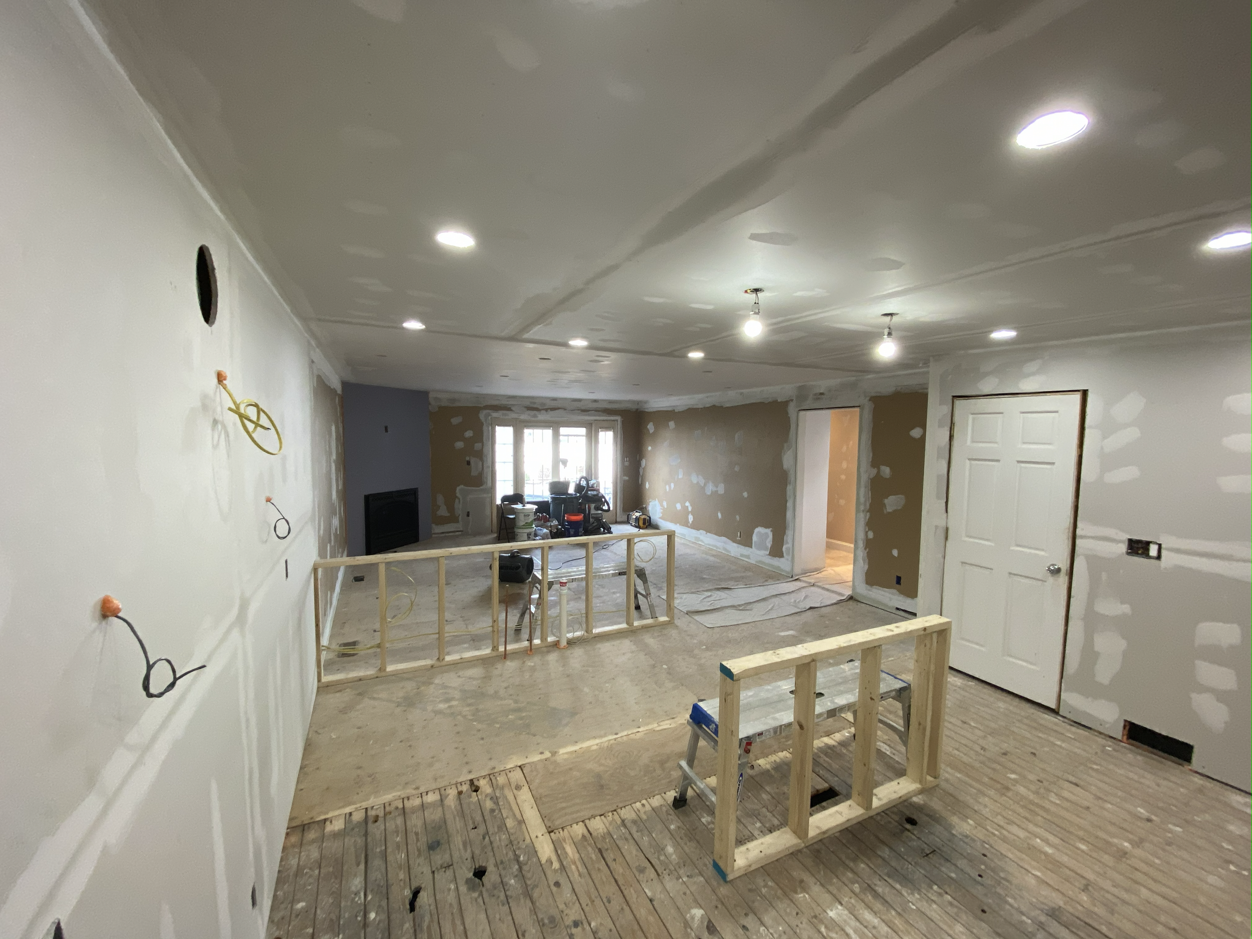 Room under renovation with drywall partially installed, electrical wiring exposed, and construction materials scattered, including a wooden frame and step ladder.