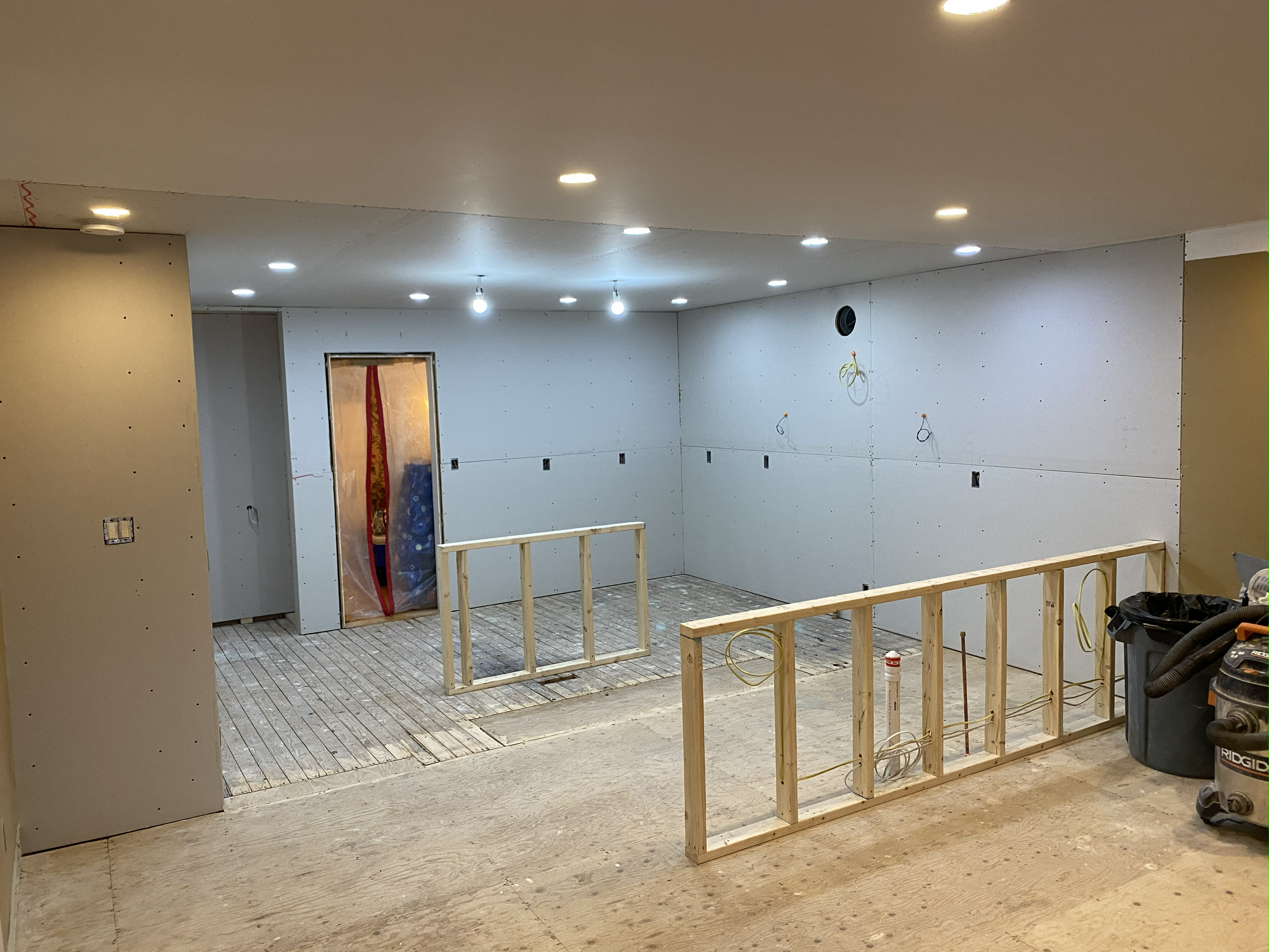 Interior of a room under construction with drywall on the walls, electrical wiring hanging, and partially built wooden framing.