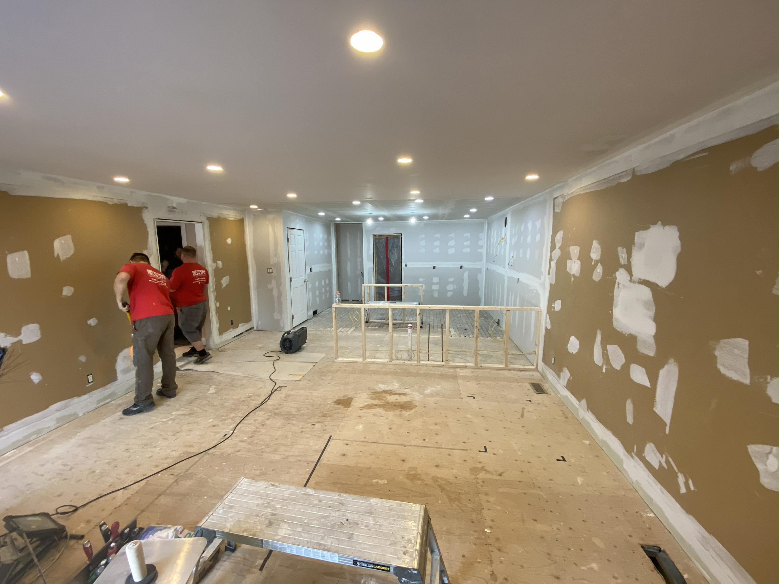 Two construction workers in red shirts working on drywall installation or finishing in a room under renovation, with drywall patches visible on the walls, a partially constructed safety barrier, and construction tools on the floor.
