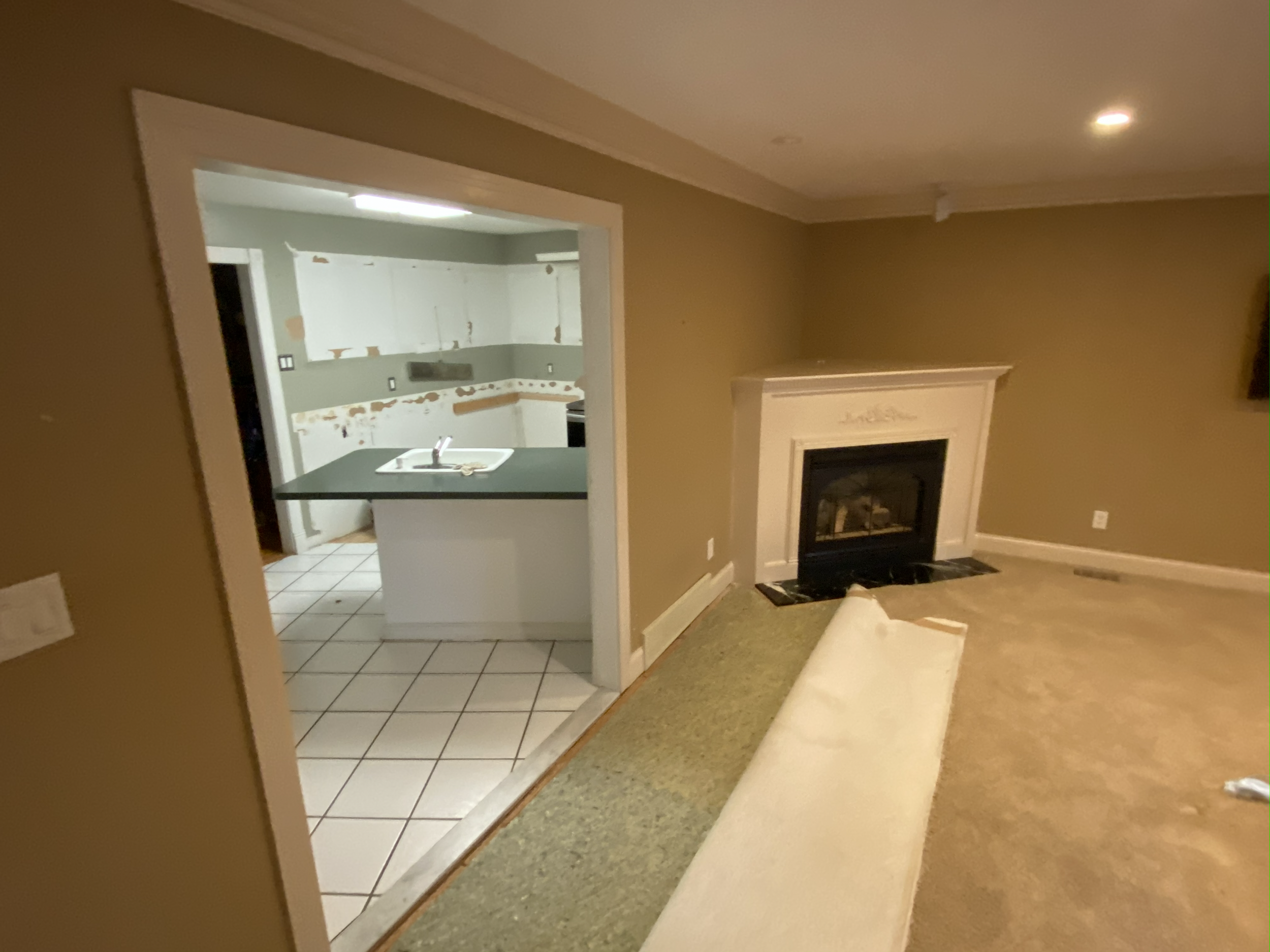 Empty living room with fireplace, beige walls, and a view into a kitchen with white cabinets and a black countertop. The kitchen appears to be under renovation or in disrepair.