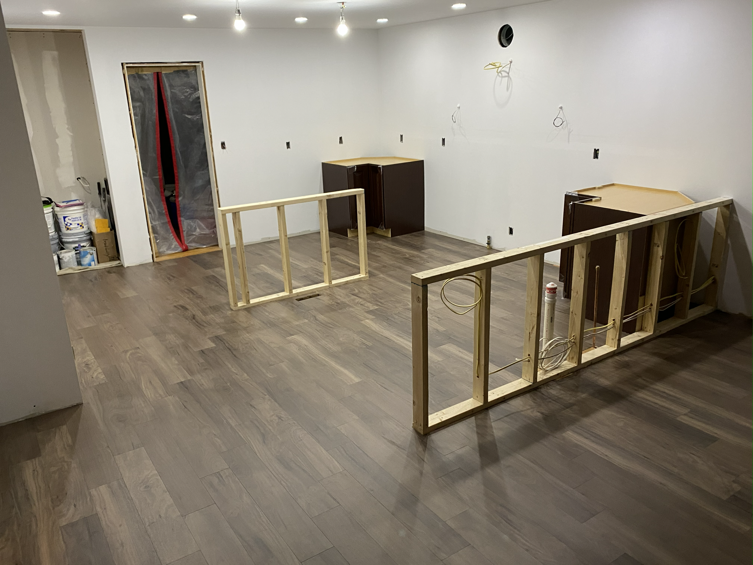 Interior of a room under renovation with partially constructed walls, exposed electrical wiring, and wooden framing, with cabinets and construction supplies along the walls.