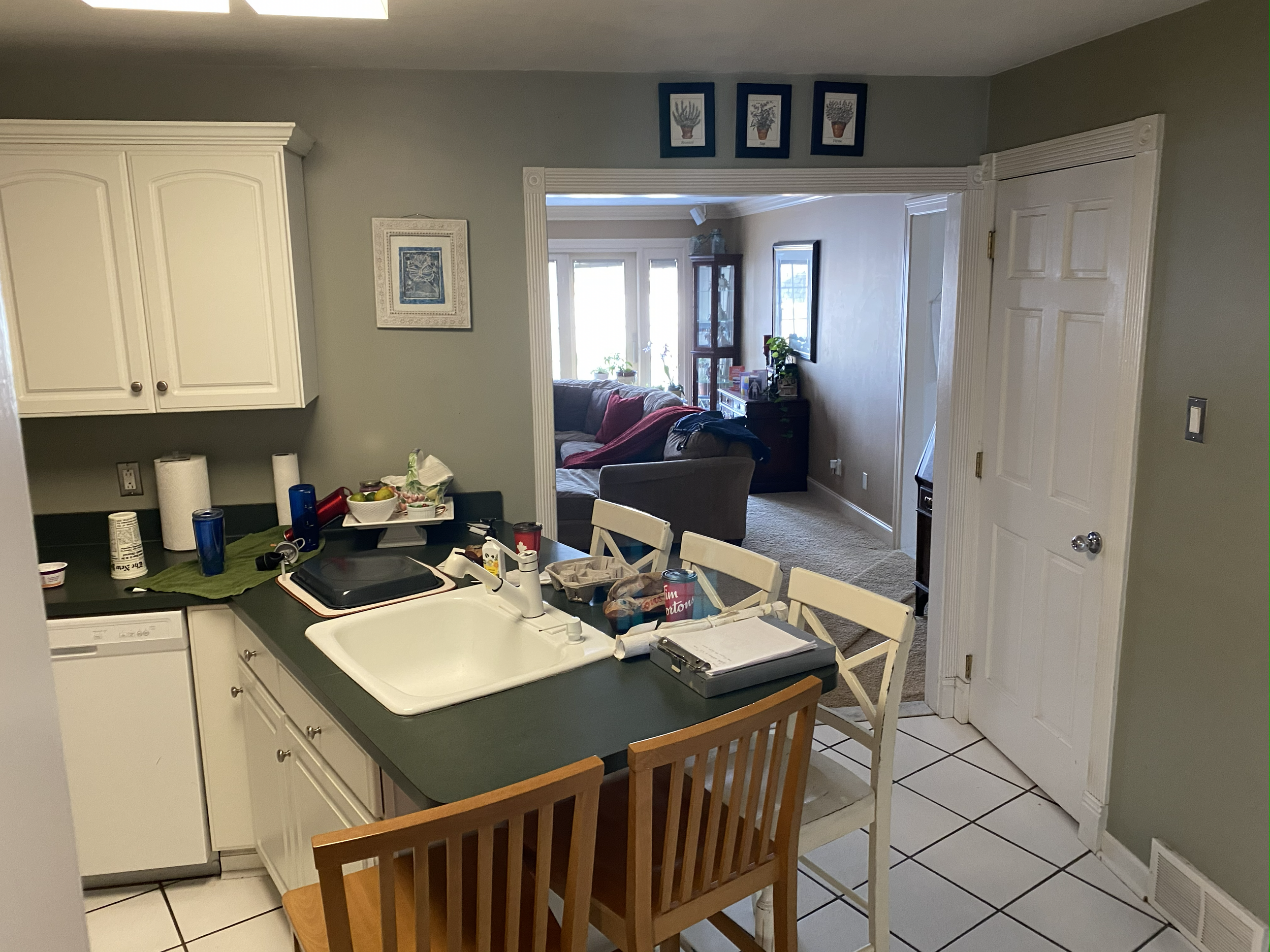 View of an open-concept kitchen and living room. In the kitchen, there are white cabinets, a green countertop, a white sink, and a small counter with various items including a blue and red water bottle, paper towels, and food containers. There's a sm
