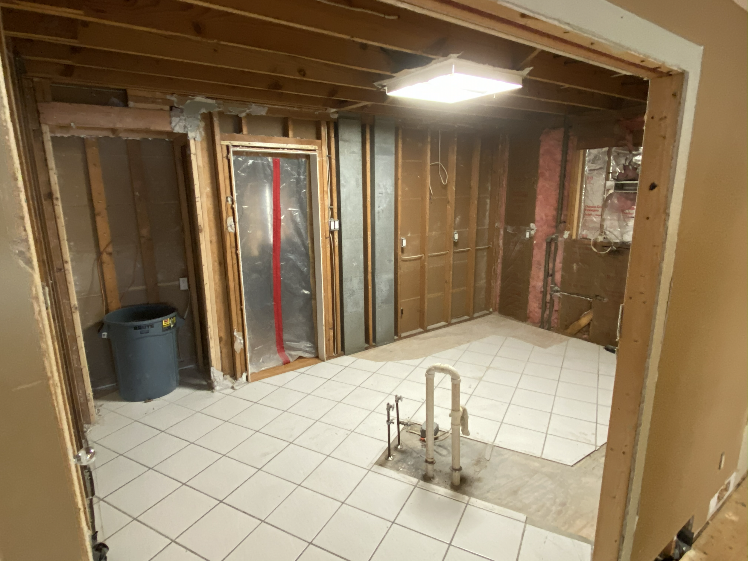 A room under renovation with exposed wooden studs, partially finished tiled floor, and a ceiling light. Construction materials and electrical wiring are visible, with a gray trash bin in the corner.