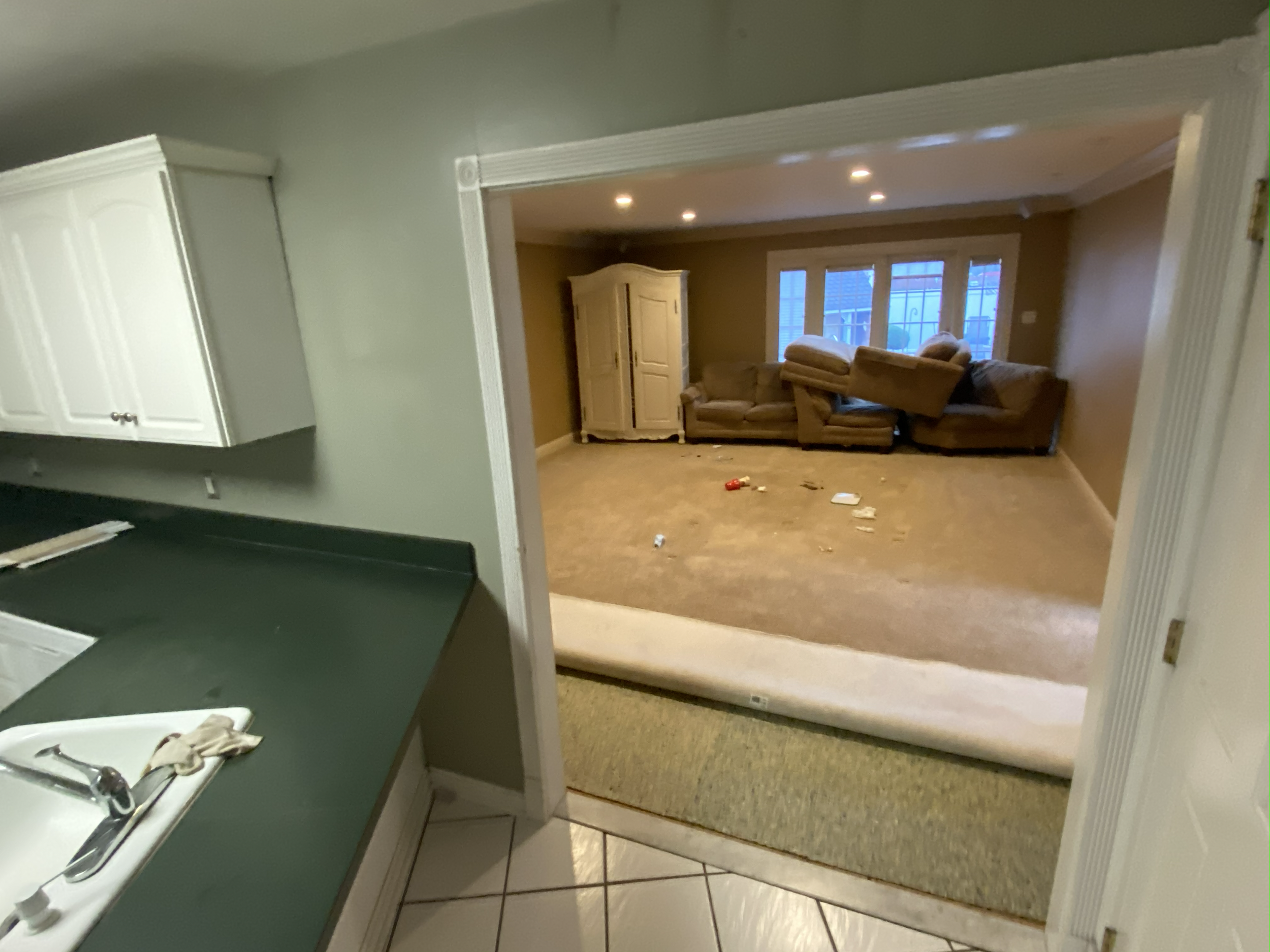 View from the kitchen showing a living room with a green countertop, white cabinets, a sink with a cloth, and a carpeted floor with scattered trash and a rolled-up rug in the open space.