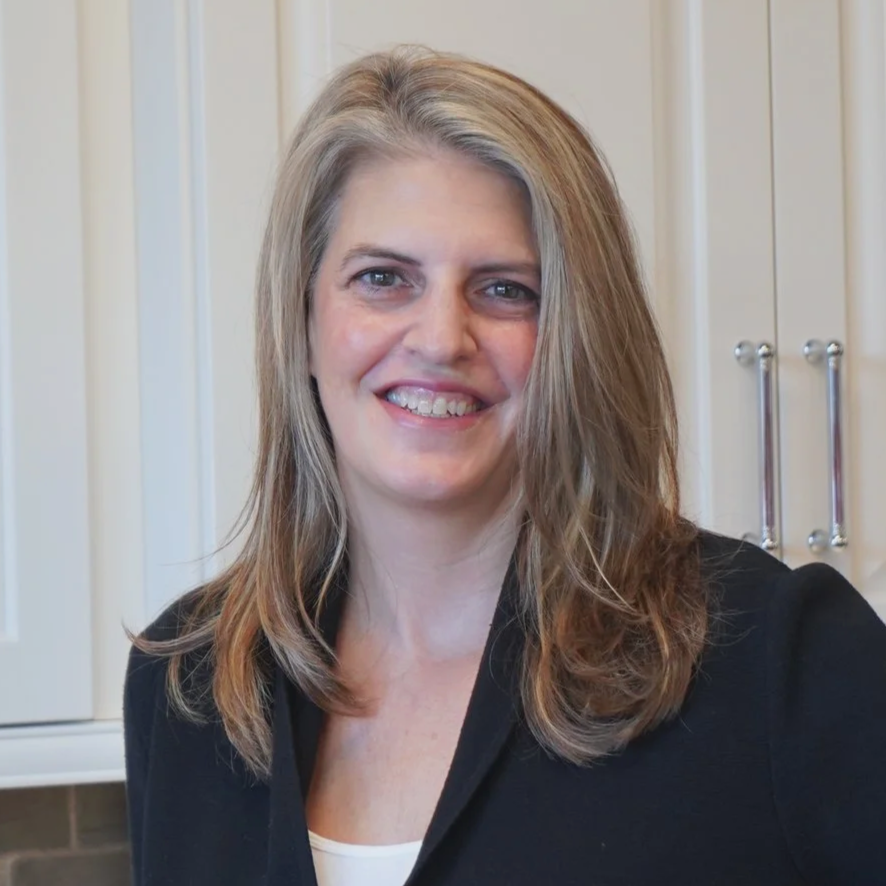 A woman with shoulder-length light brown hair smiling in a kitchen with white cabinets.