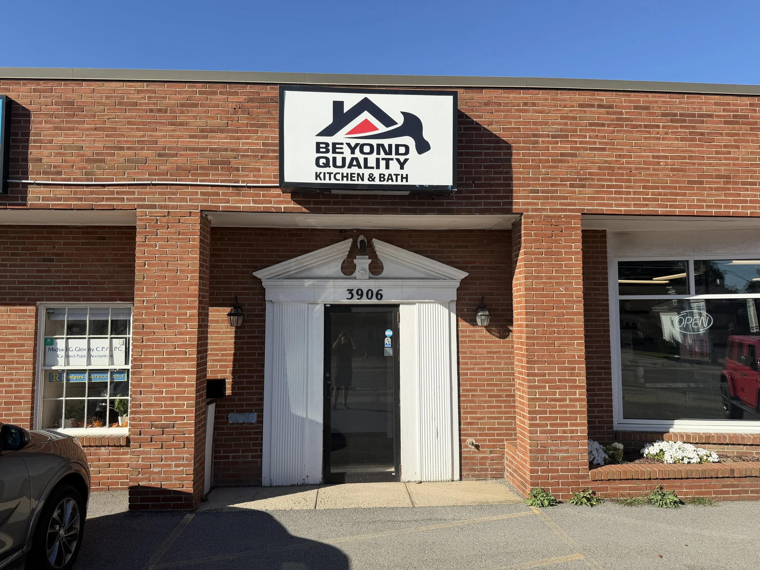 Brick storefront with a sign that reads 'Beyond Quality Kitchen & Bath', a white entrance door with the number 3906 above it, and a window displaying an 'Open' sign and a red vehicle outside.