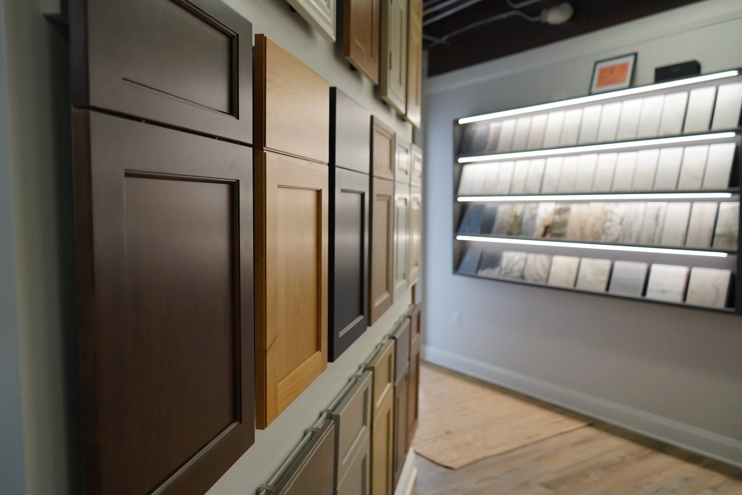 Display of various cabinet door samples in different colors and finishes at a flooring or home improvement store, with a wall of tile samples and a curtain of natural light in the background.