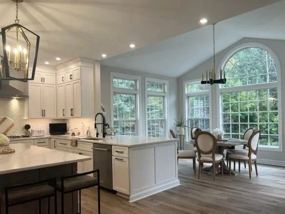 Bright open kitchen and dining area with white cabinets, a central island, and large windows with a view of greenery.
