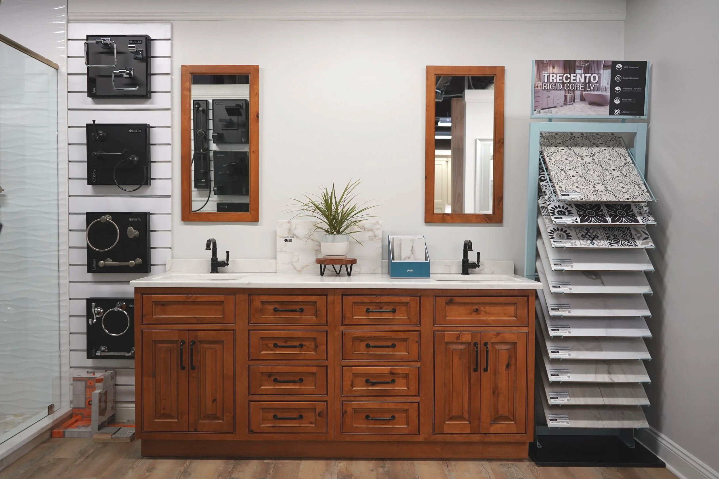 Bathroom vanity with two sinks, a wooden cabinet with multiple drawers and doors, two mirrors with wooden frames, a potted plant, and a display rack with tile samples on the right.