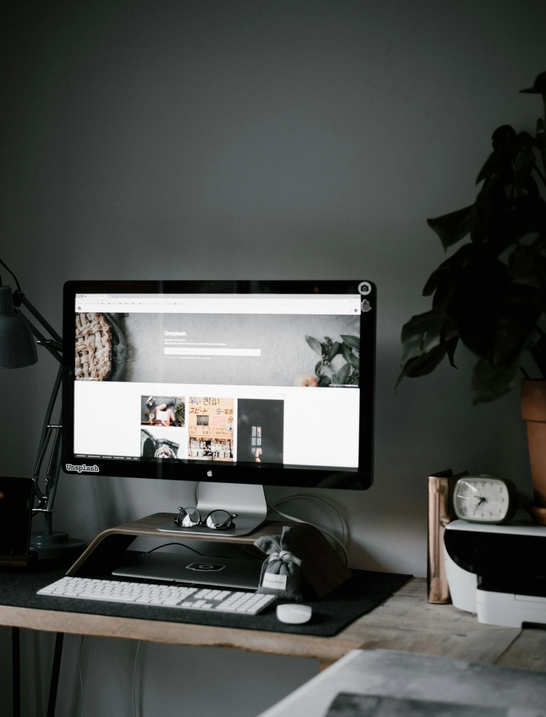 Photo of a workspace with a desktop computer, keyboard, mouse, glasses, a closed laptop, a desk lamp, a clock, and potted plants on a wooden desk against a plain wall.
