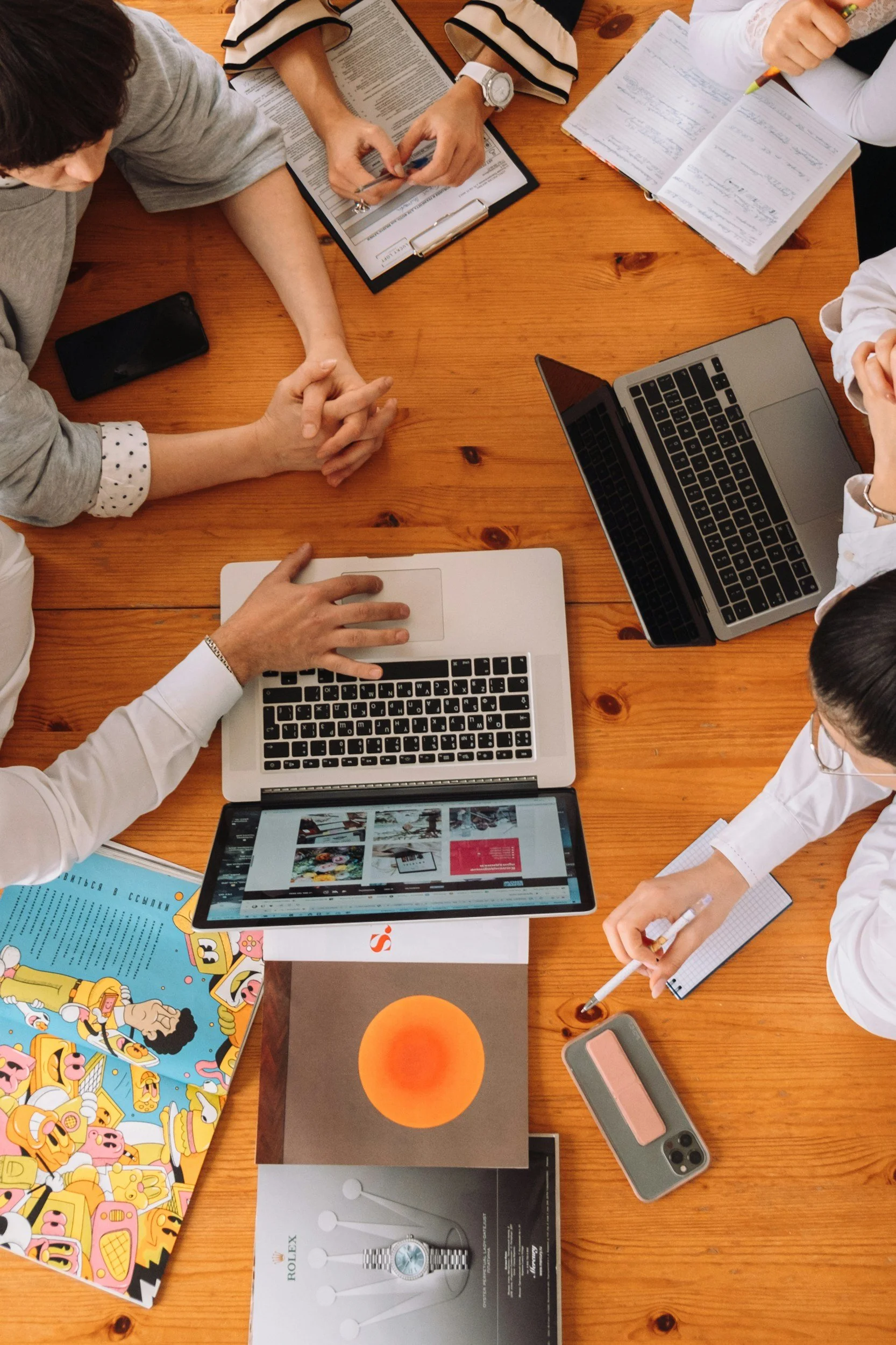A top-down view of a wooden table with several laptops, notebooks, books, a smartphone, and a few people working or studying together.