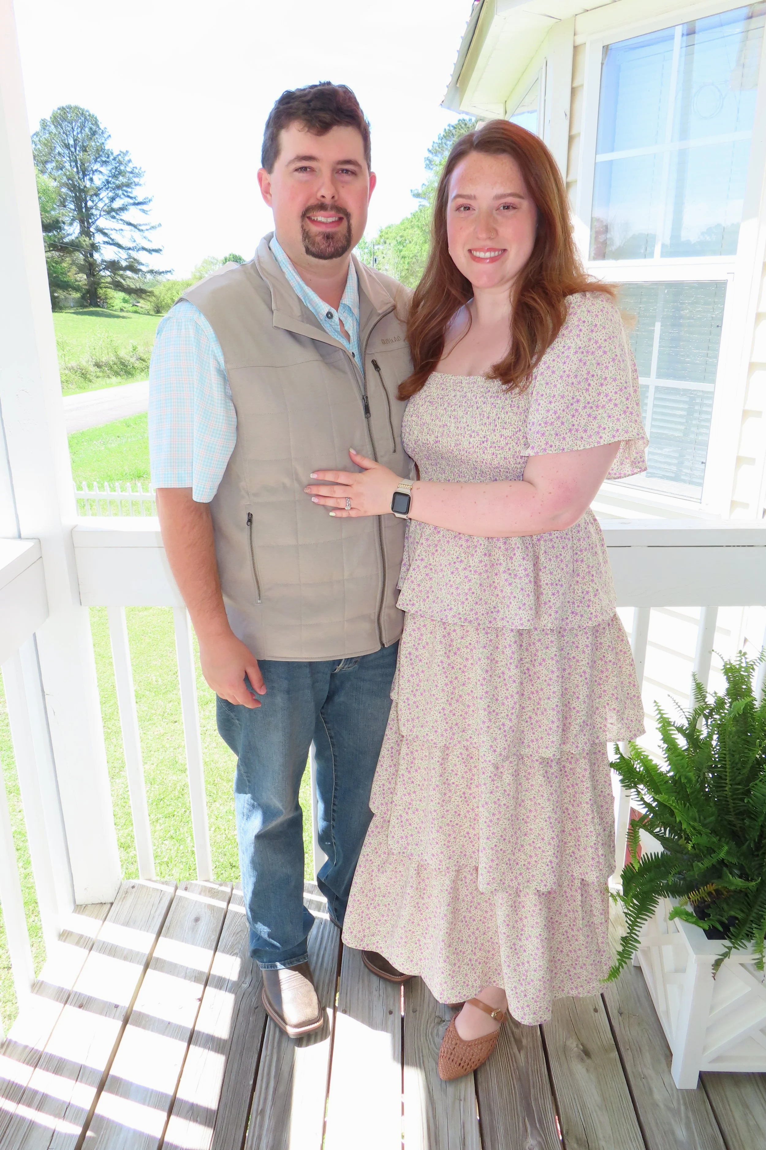 A man and woman standing close together on a porch, smiling at the camera, with trees and a bright sky in the background. The woman is wearing a floral dress and brown shoes, and the man is wearing a light shirt, vest, jeans, and brown shoes