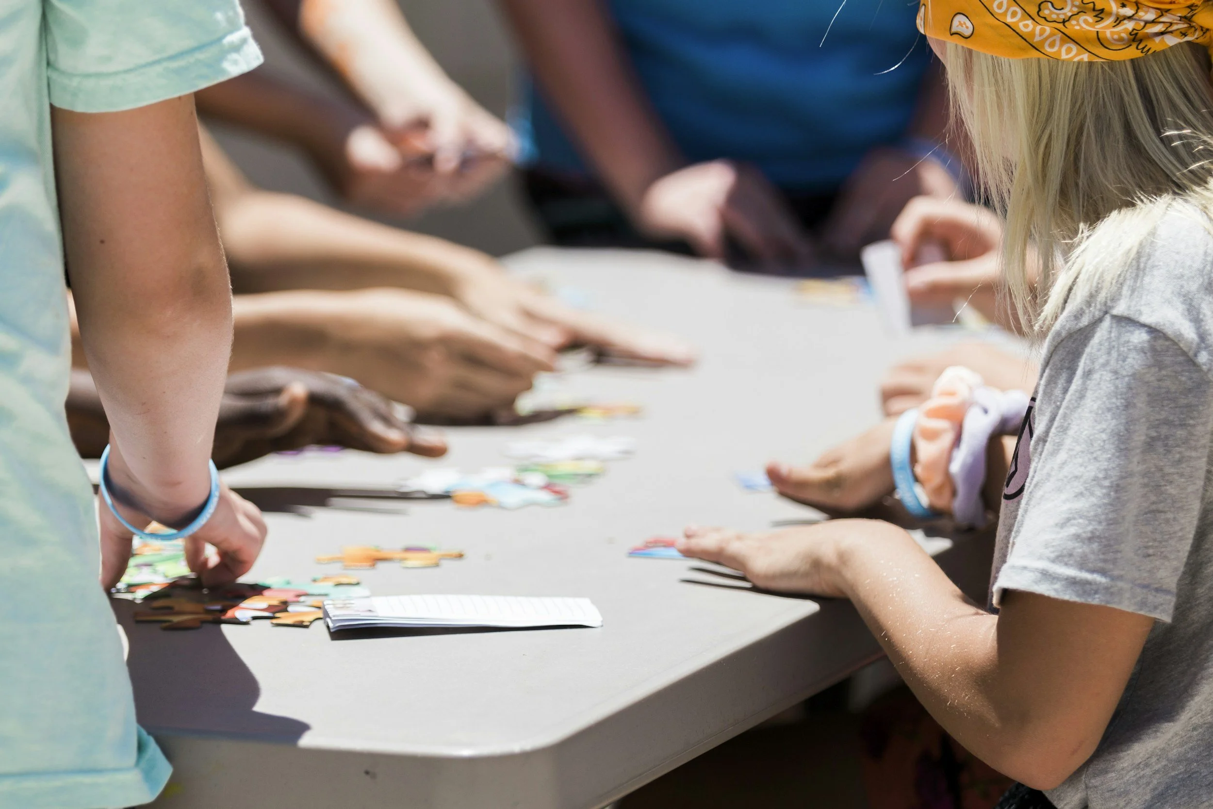 Children gathered around a table playing a card game outdoors in the sunlight.