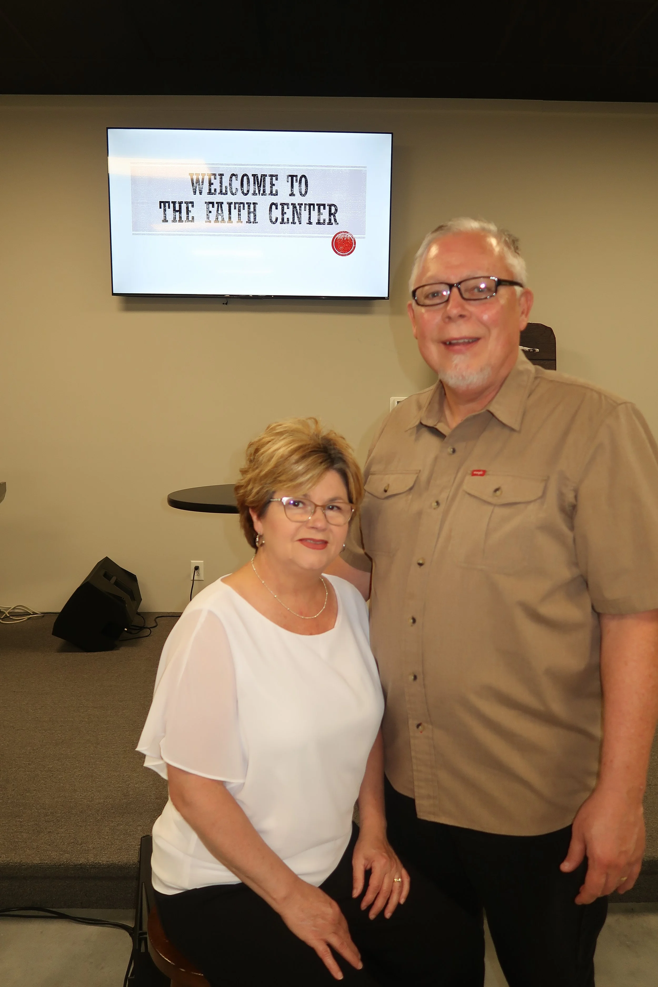 A woman with short brown hair and glasses wearing a white blouse and a man with short gray hair, glasses, and a tan shirt standing together in a room with a sign that reads "Welcome to The Faith Center" on a TV screen in the background.