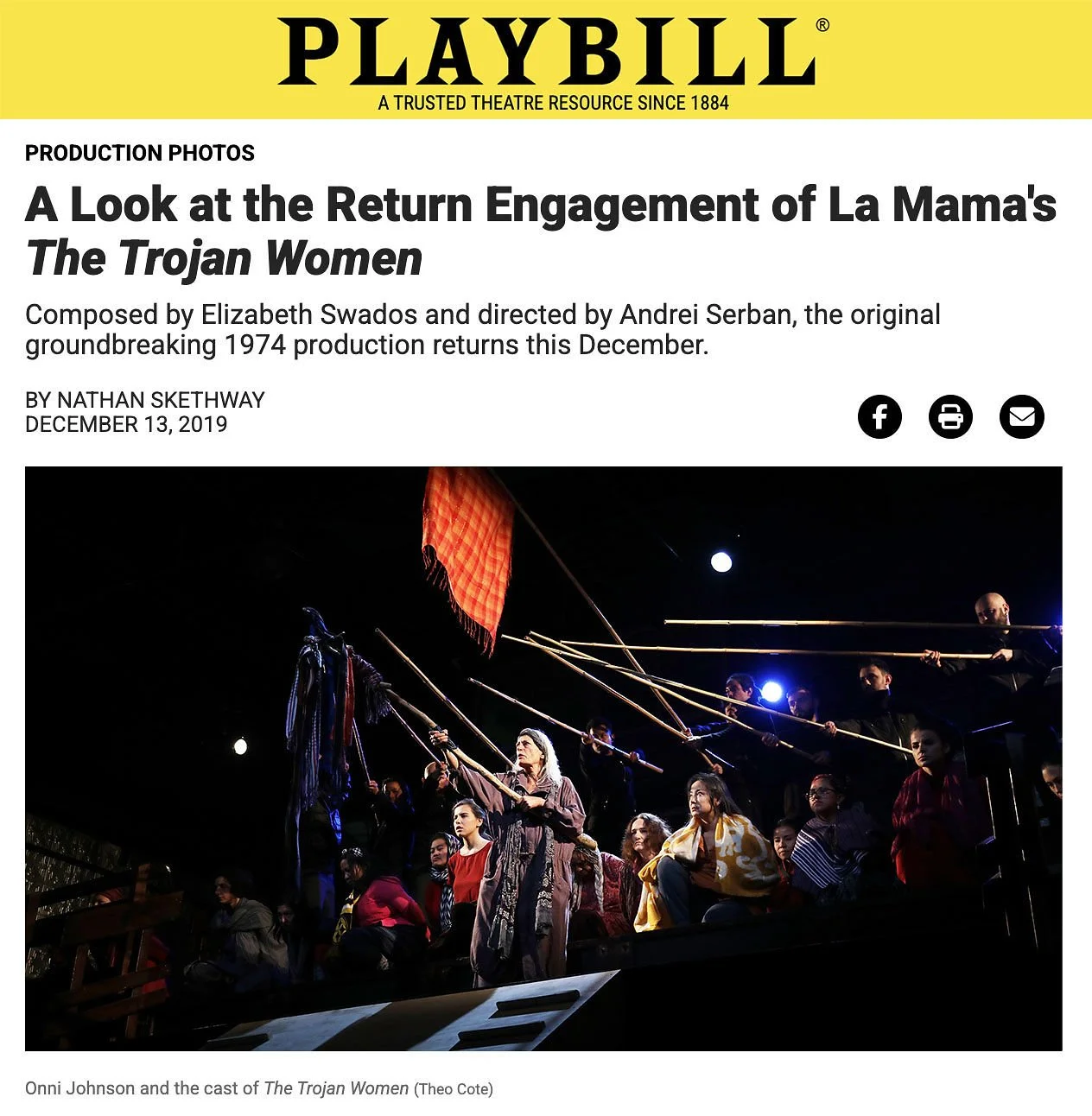 The photograph shows a theatrical scene with cast members of La Mēna's The Trojan Women. One central actress is standing, holding a flag, amid other actors seated and standing around her in dramatic poses. The stage is dark with spotlights highlighti