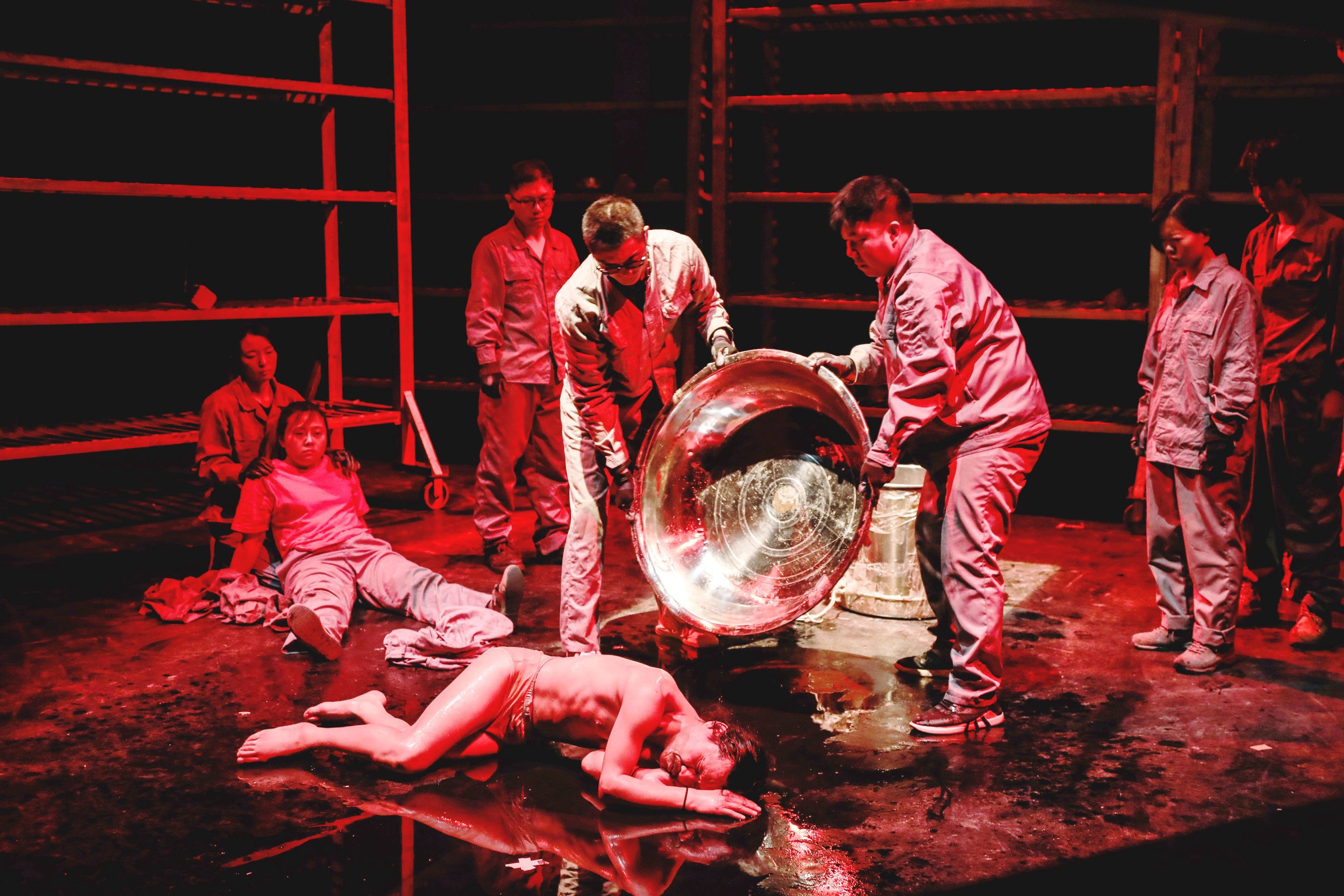 The image depicts a theatrical or performance scene with multiple actors in costumes, some sitting and others standing, around a large metallic object. The lighting is red, and the setting appears to be an industrial or warehouse-like space with shel