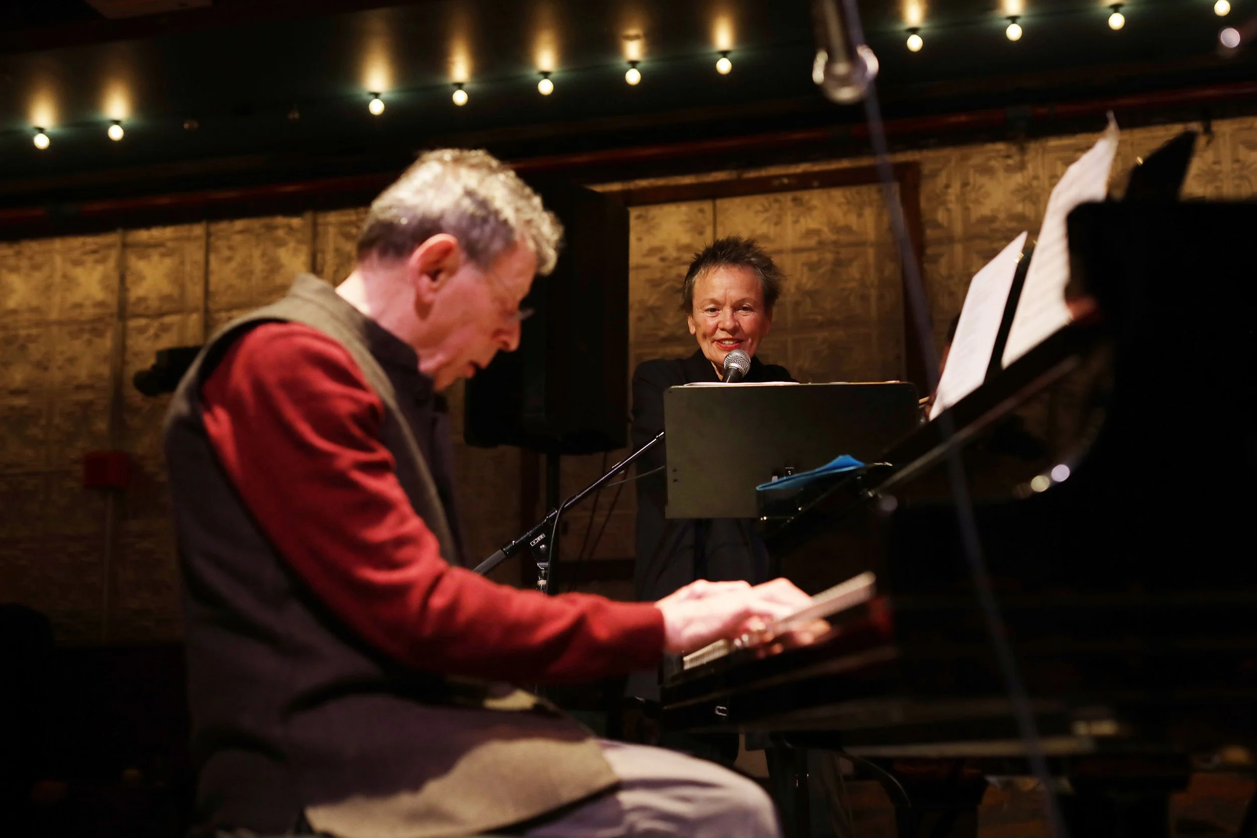 A woman singing into a microphone while playing the piano, with a man focused on playing the piano in the foreground.