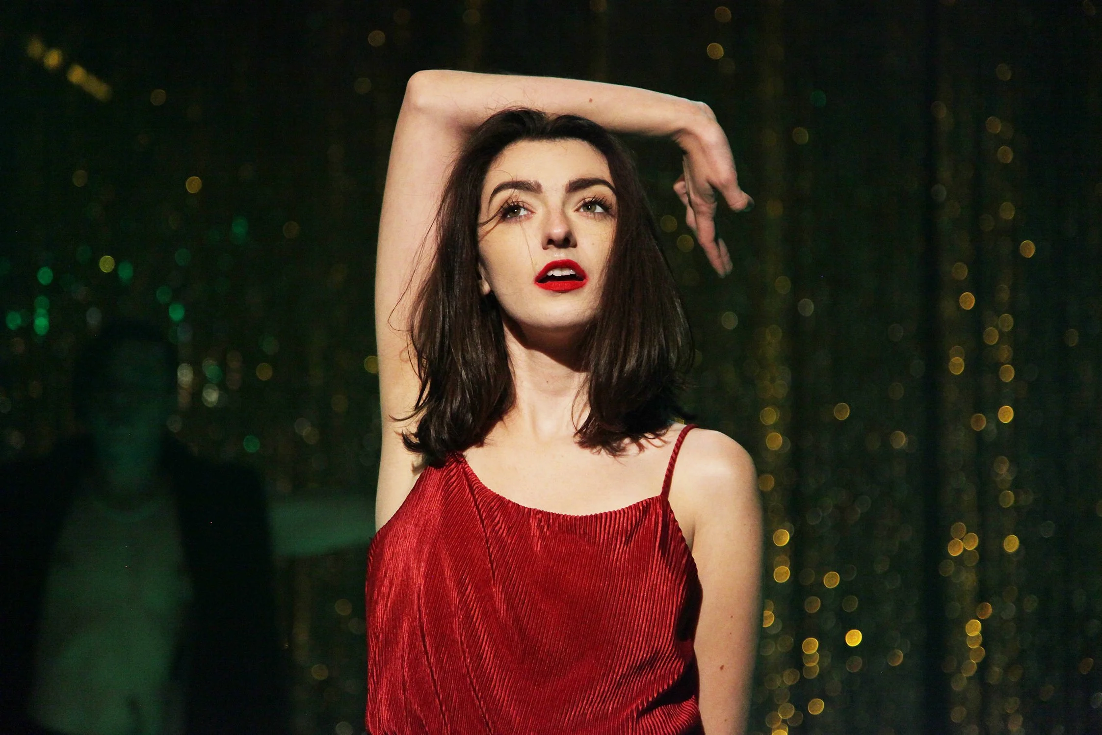 A woman with dark brown hair wearing a red dress poses with her right arm raised above her head against a dark background with blurred lights.