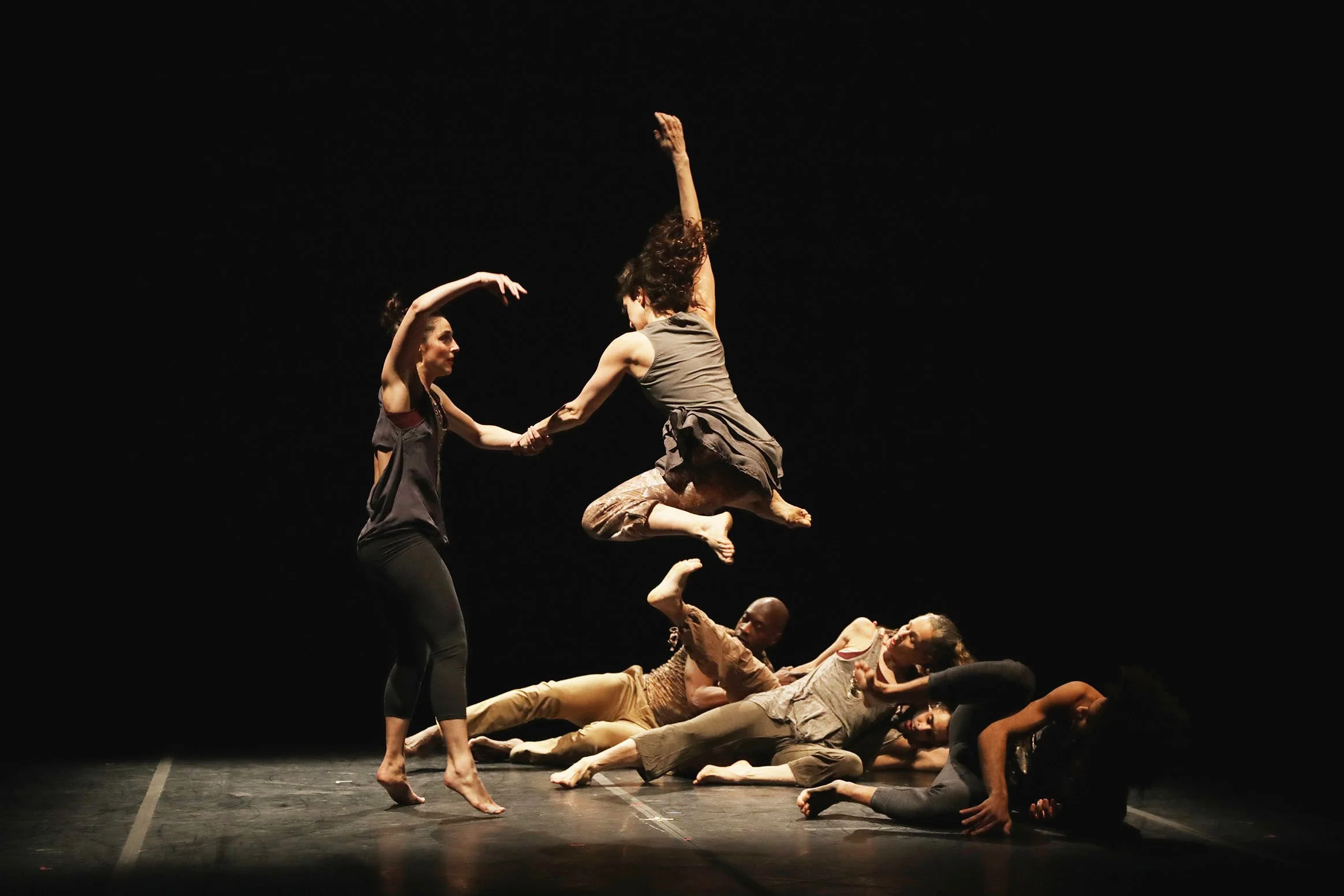 Dancers perform on a dark stage, with one dancer mid-air and others on the ground holding handles of the airborne dancer.