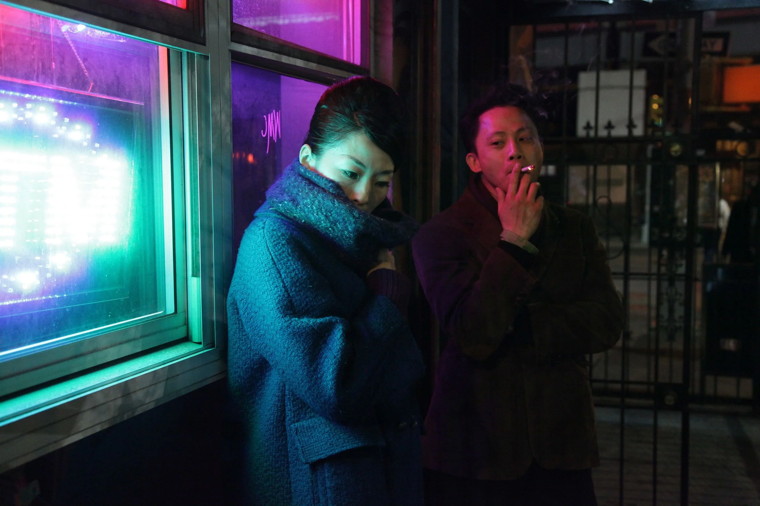 Two people, a woman and a man, standing outside at night near a neon-lit window. The woman has her arms crossed, looking down, and the man is smoking a cigarette while looking at her. The scene is illuminated by pink and purple neon lights.