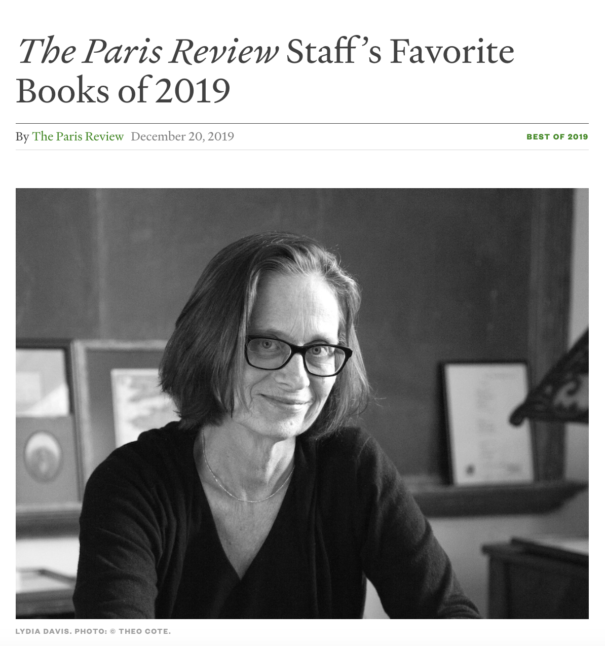 Black and white photo of Lydia Davis sitting at a desk, smiling, wearing glasses and a dark top, with a framed document and artwork in the background.