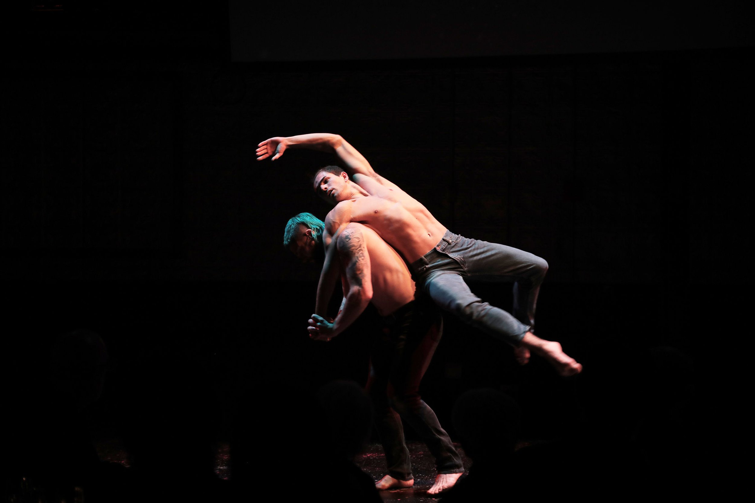 Two shirtless male dancers perform a contemporary dance routine on a dark stage, with one dancer bending backward over the other's back as the other supports him. One dancer has teal hair tattoos, and both are focused and expressive.