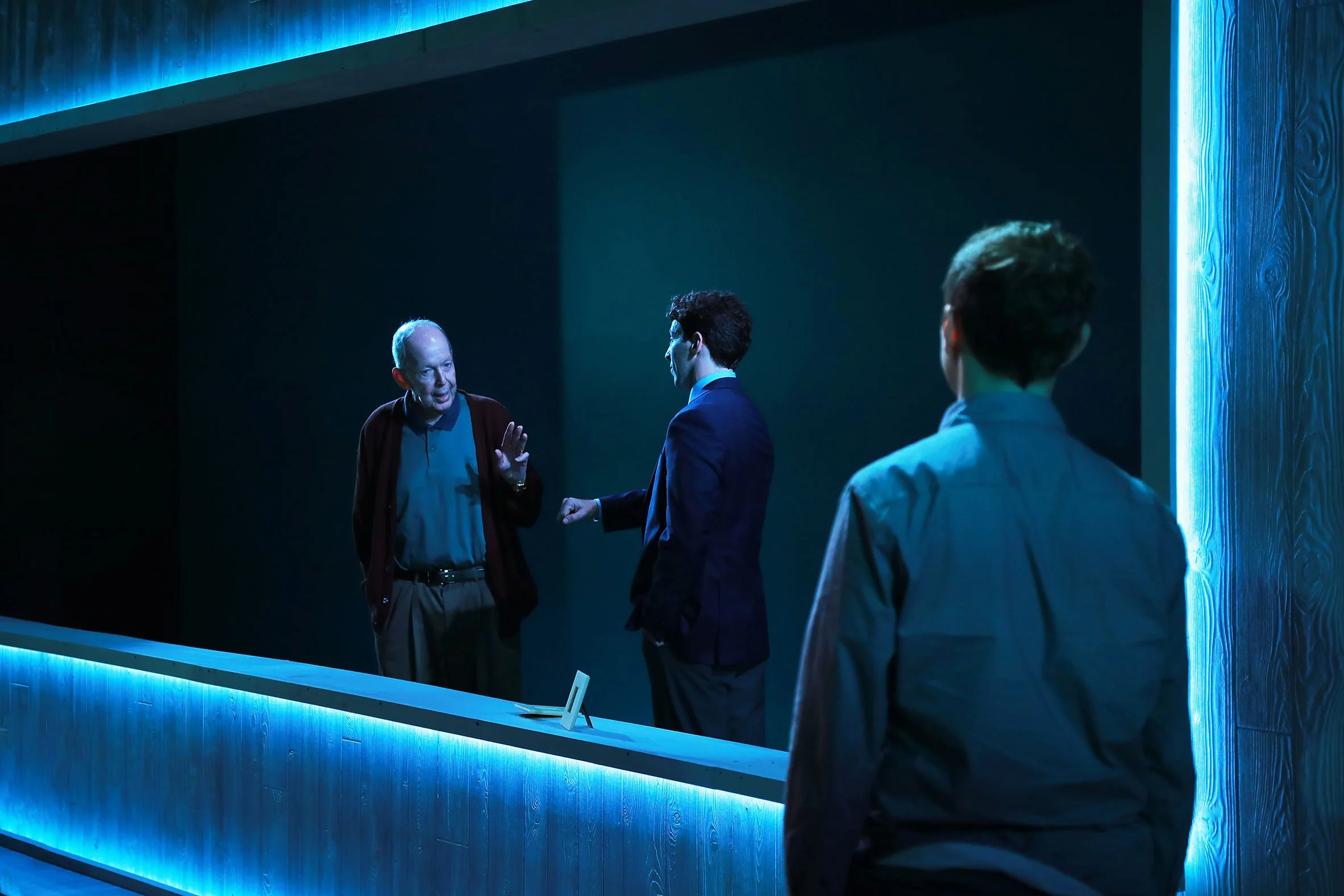 Three men engaged in conversation behind a reception desk illuminated with blue lighting.